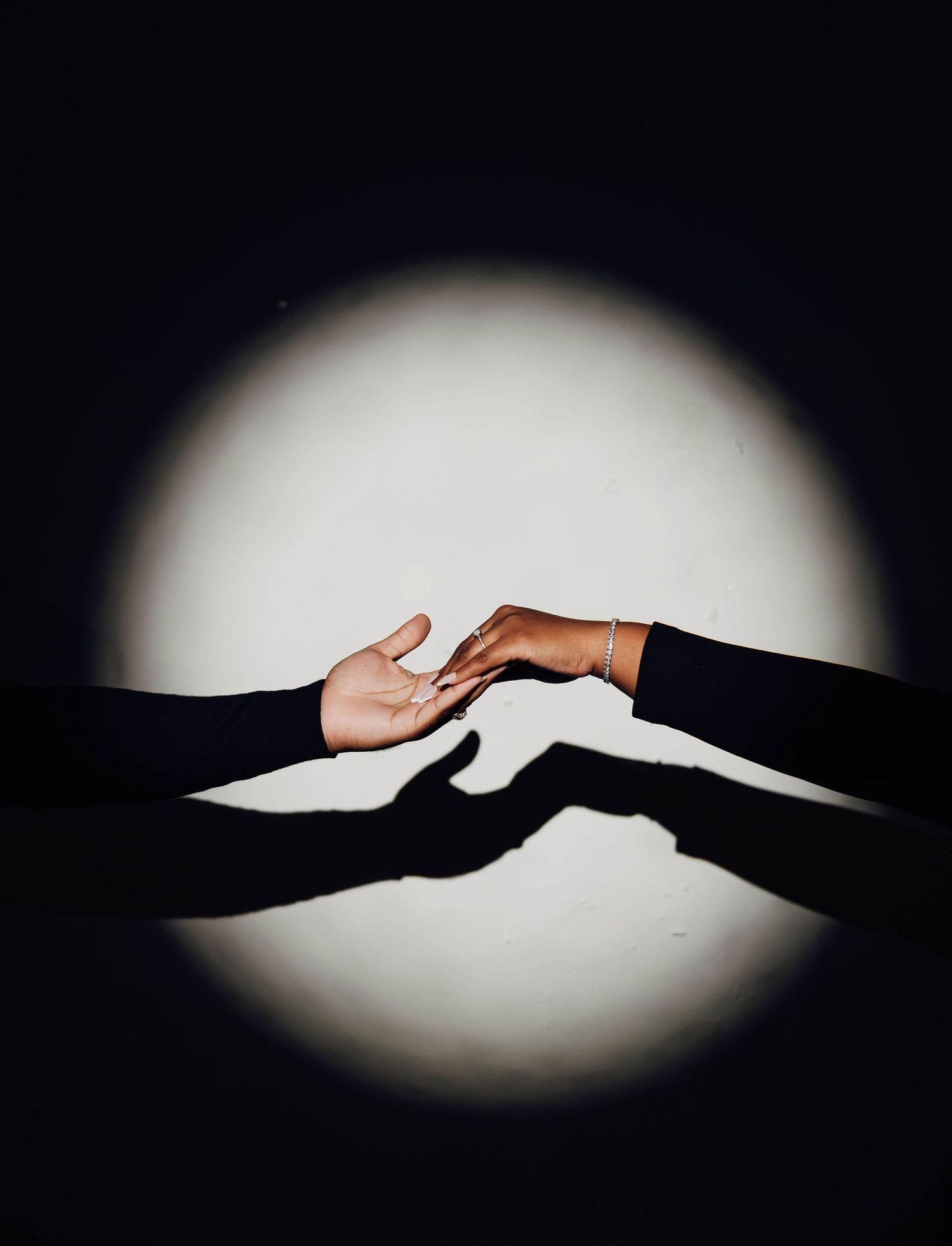 Two hands reaching toward each other in a spotlight against a dark background, casting dramatic shadows.