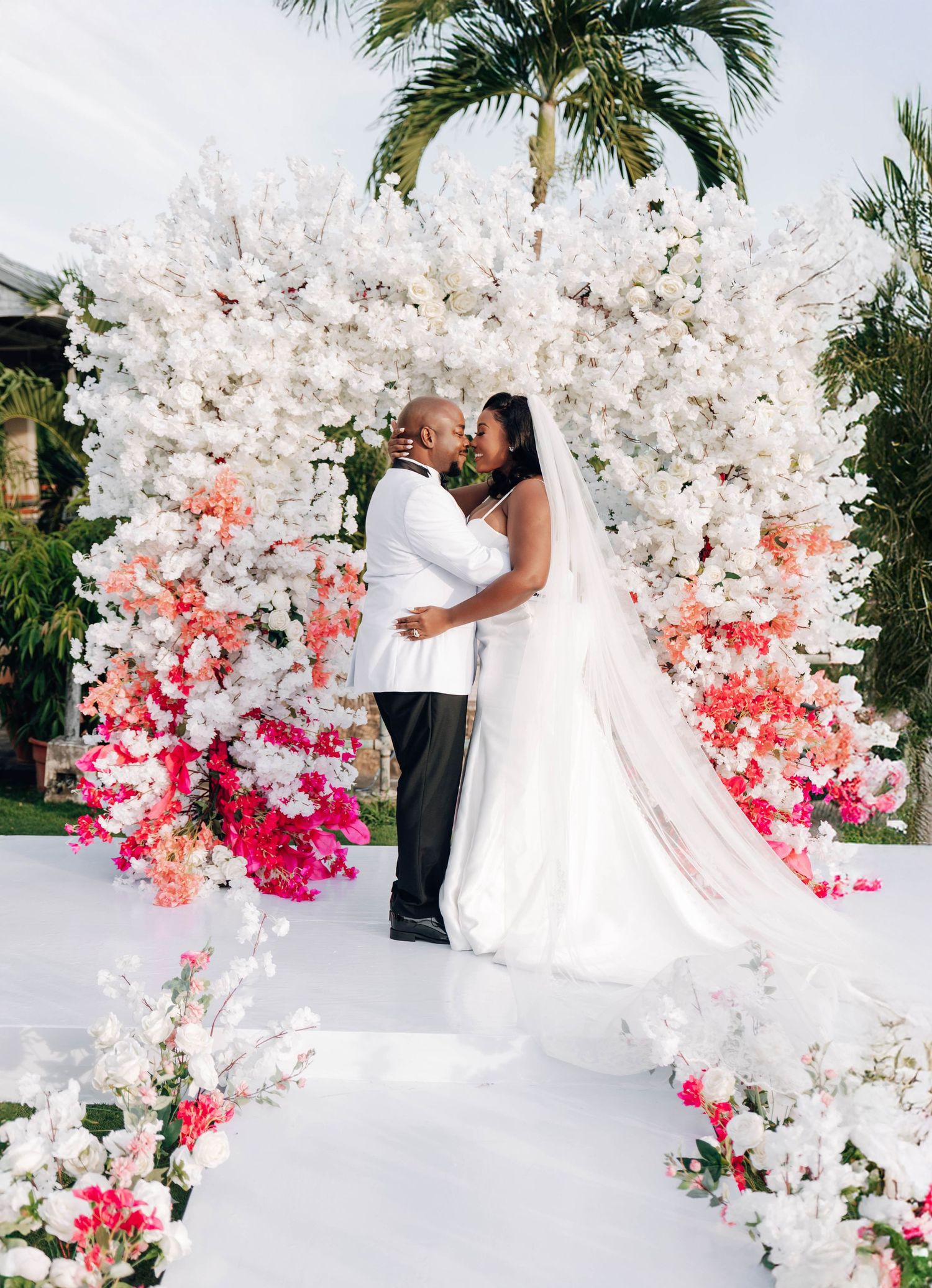 Couple shares first kiss at outdoor wedding ceremony beneath stunning white and pink floral arch with palm trees.
