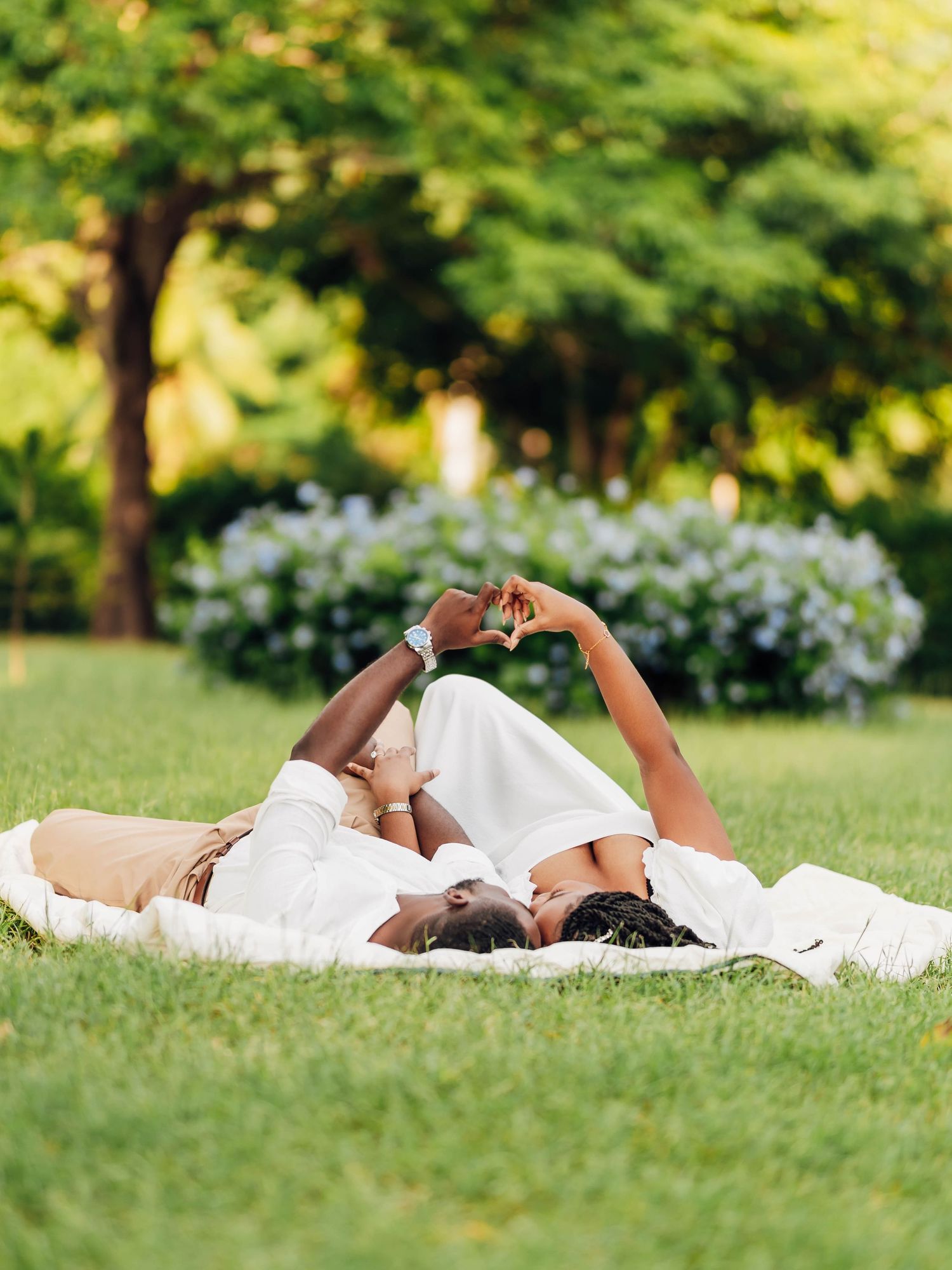 Couple lying on grass in a park, making a heart shape with their hands against a lush green backdrop.