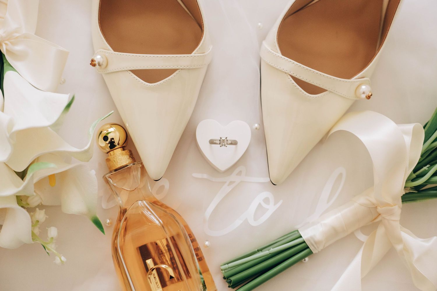 Elegant bridal flat lay with ivory pointed-toe heels, pearl ring box, white calla lily bouquet, and gold perfume bottle.