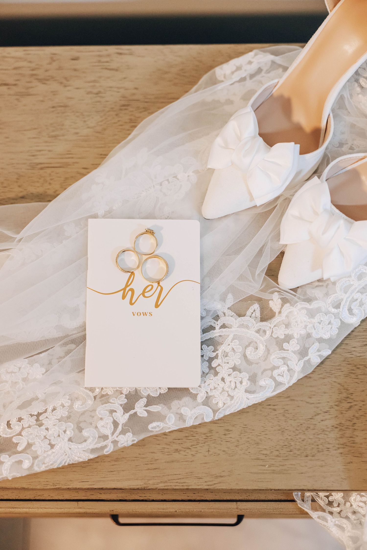 White bow heels and gold wedding rings on vow booklet atop lace bridal veil on wooden dresser.