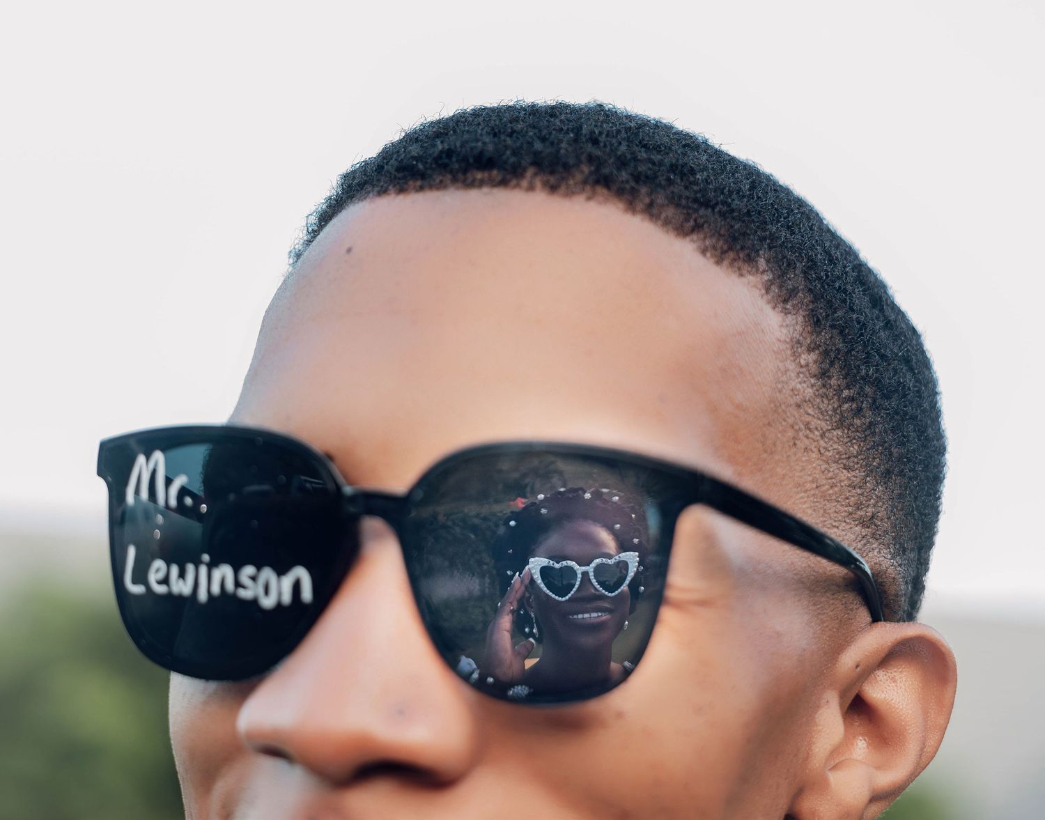Person wearing black sunglasses with Mr. Levinson written on the lens, reflecting heart-shaped glasses.
