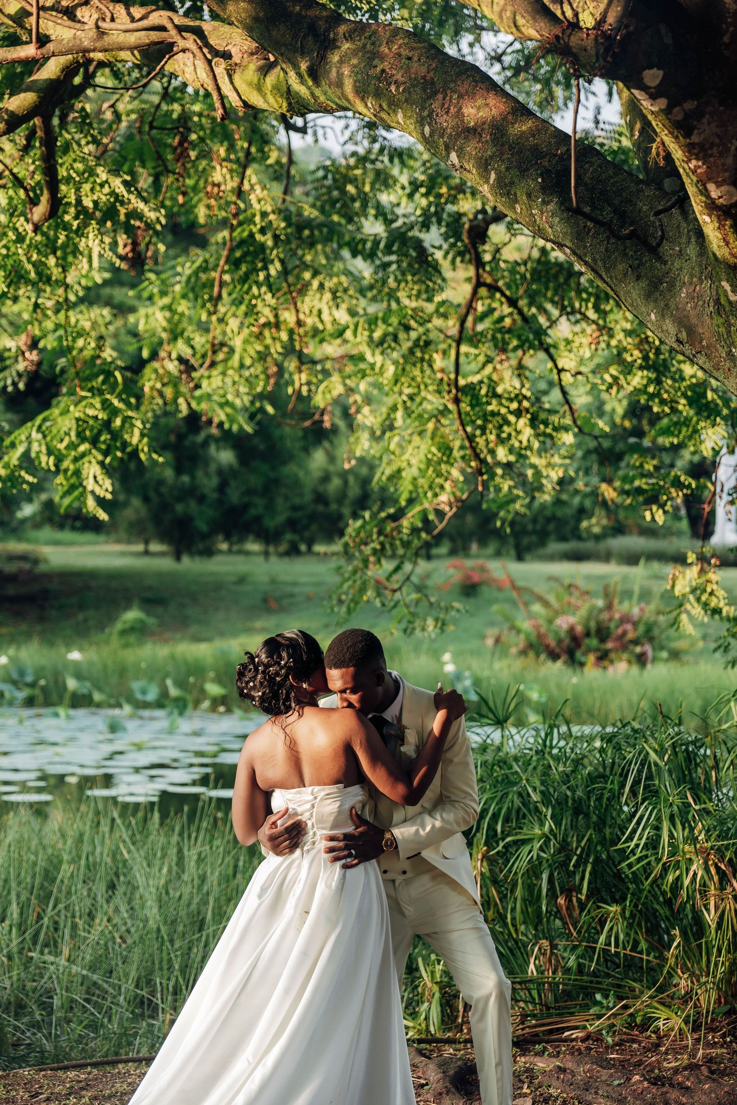 A couple shares a romantic kiss under a lush tree by a serene pond, bride in white gown, groom in cream suit.