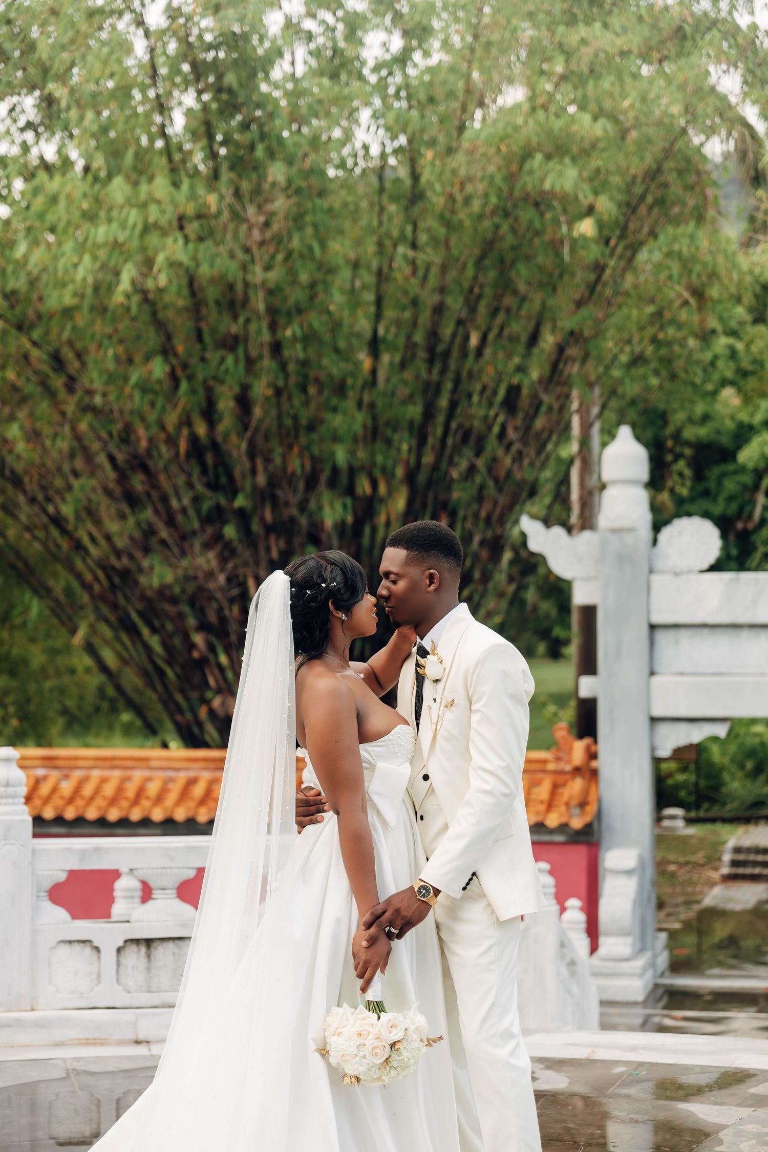Black bride and groom nearly kissing in a garden, both dressed in white, with Asian-inspired architecture in the background.