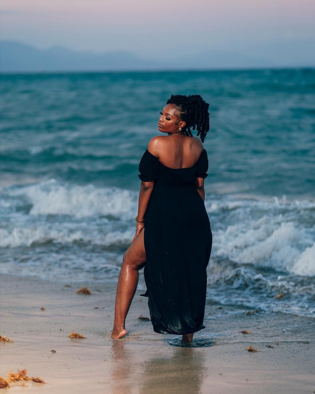 Woman in black off-shoulder maxi dress standing barefoot on a beach with ocean waves at dusk.