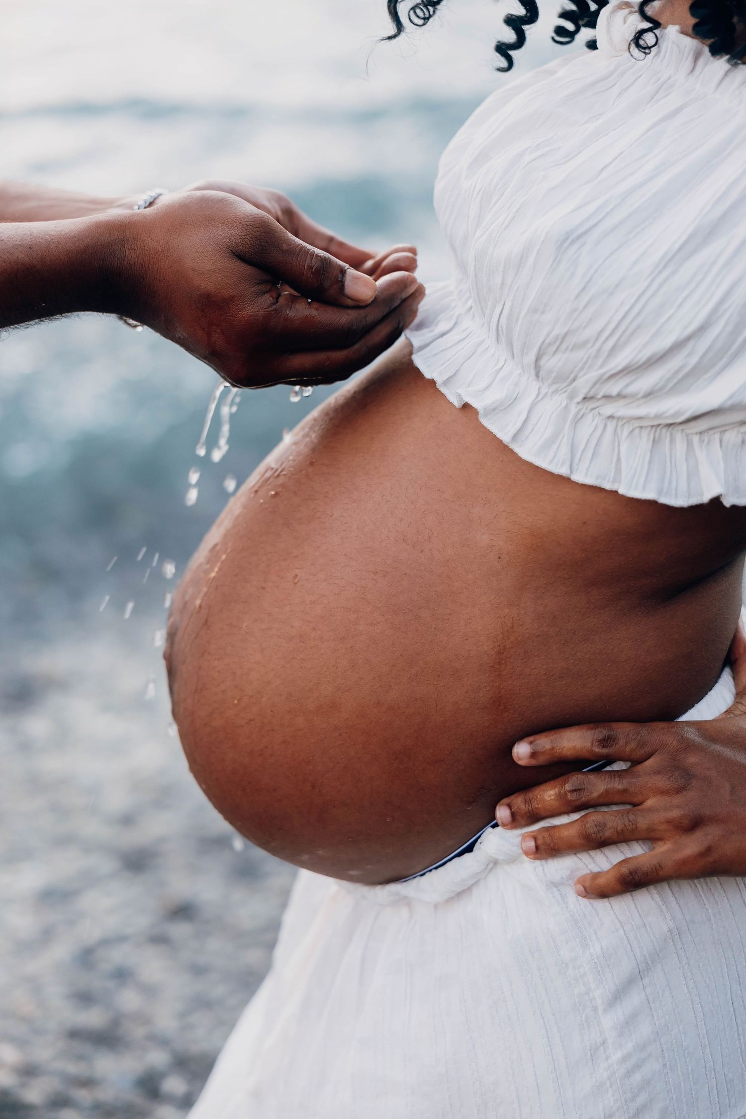 A pregnant woman in a white dress has water poured over her bare baby bump near a serene body of water.