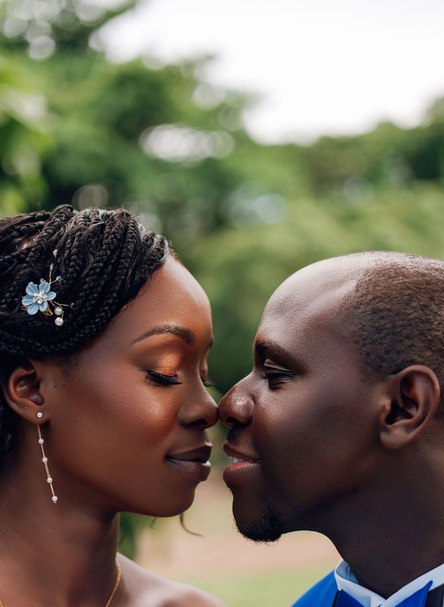 A couple shares a romantic nose-to-nose moment outdoors, bride wearing braids with a blue floral hair accessory.