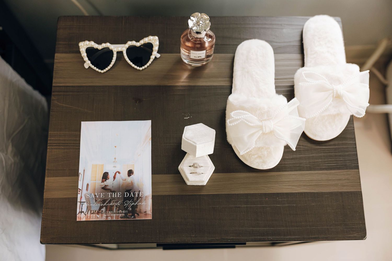 Flat lay of heart sunglasses, perfume, white bow slippers, ring box, and save the date card on a wooden table.