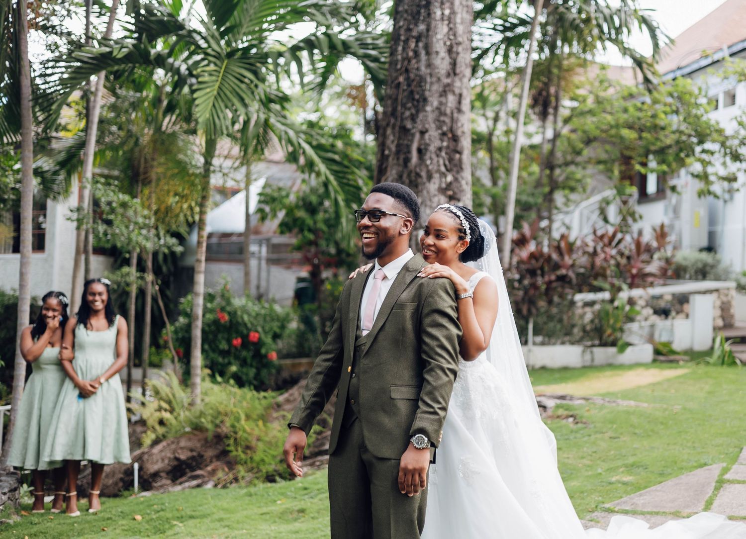 A bride and groom share a joyful first look moment outdoors, with bridesmaids in sage green dresses nearby.