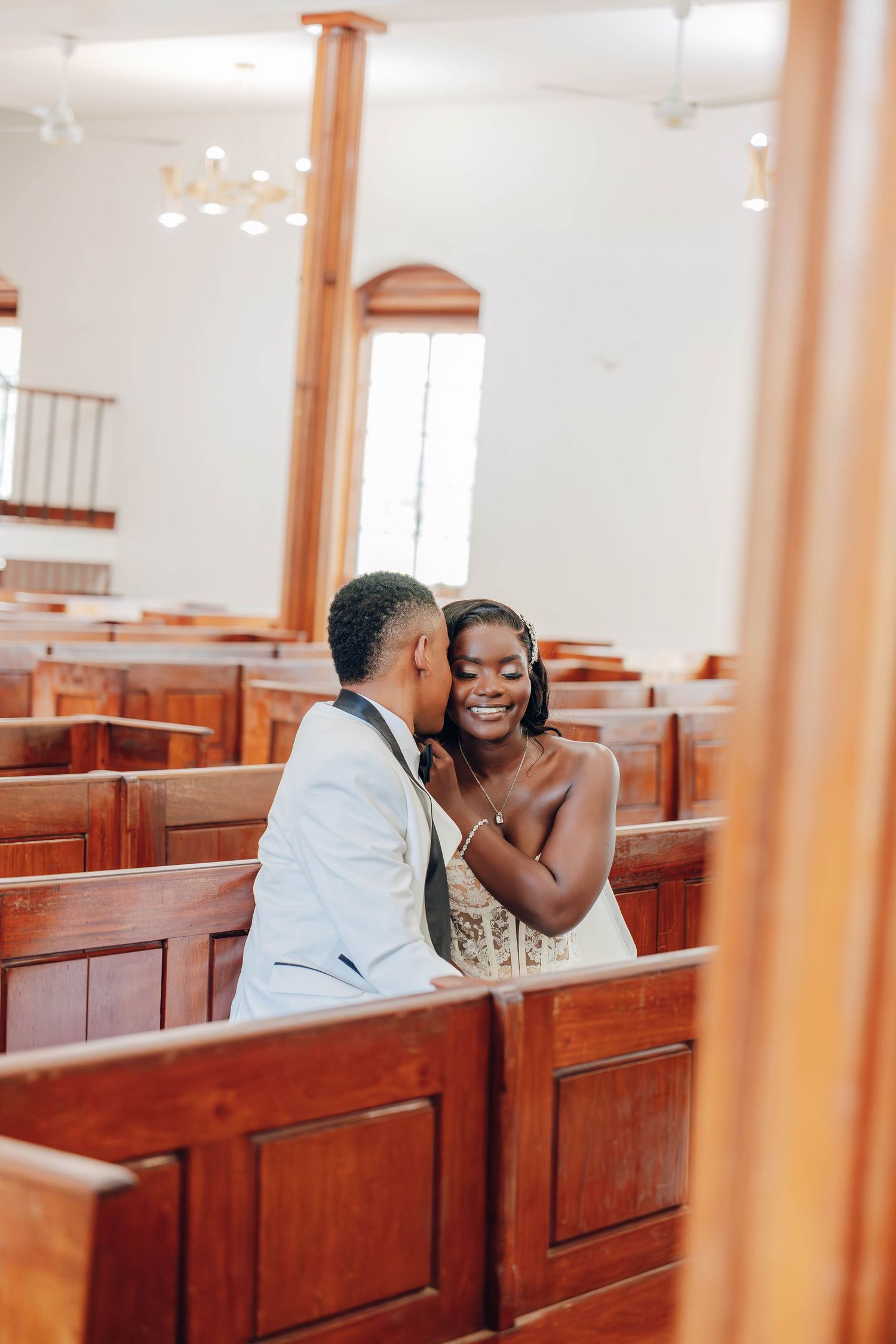 Couple sharing a romantic moment in wooden church pews, bride in white dress smiling as groom kisses her cheek.