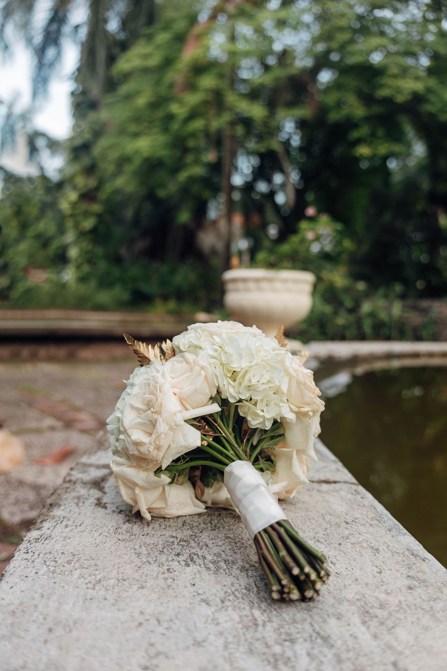 Elegant bridal bouquet of white roses and hydrangeas with ribbon wrap resting on stone ledge in lush garden setting.