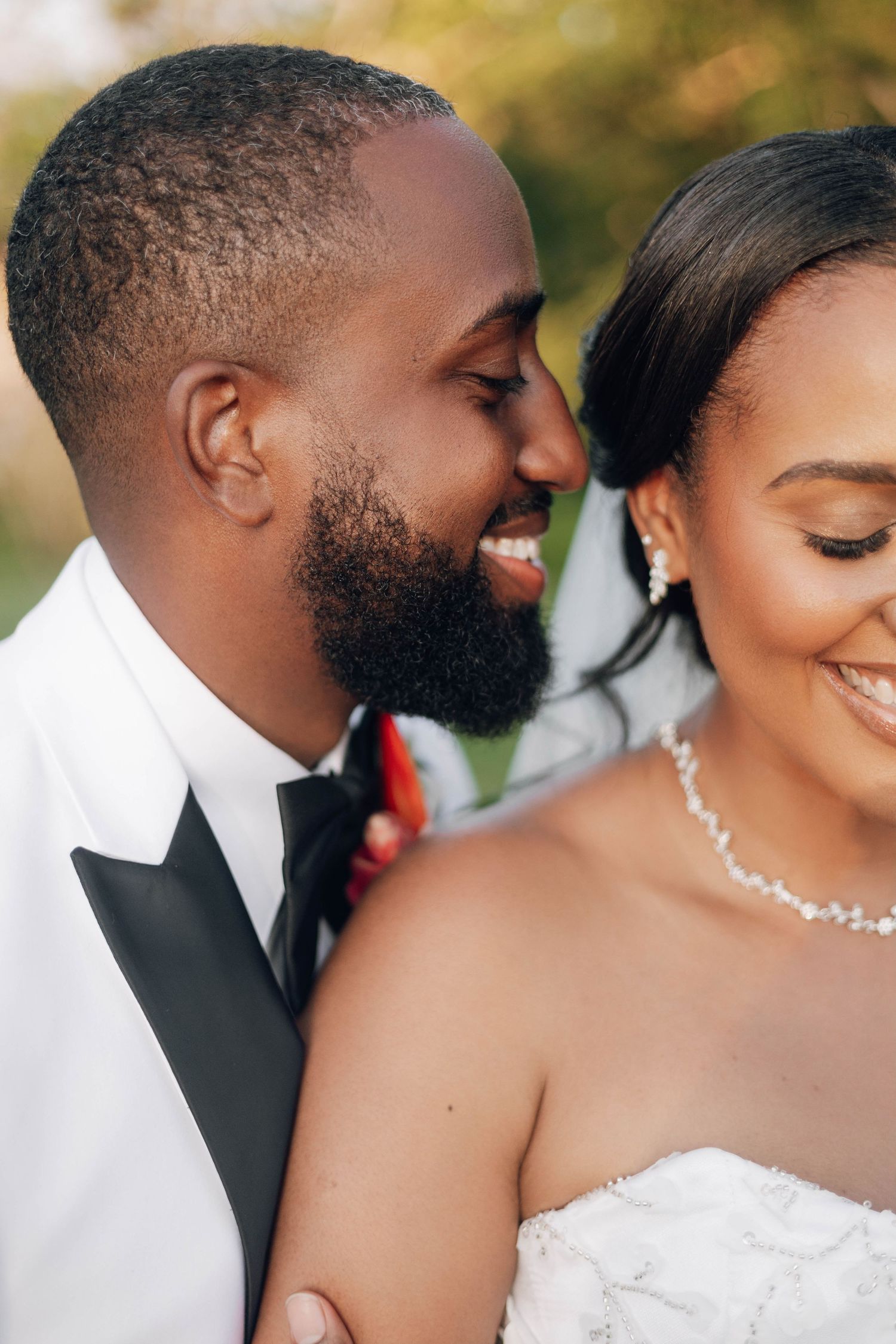 A joyful Black couple in wedding attire share a romantic moment, groom in white tuxedo, bride in white gown with pearl necklace.