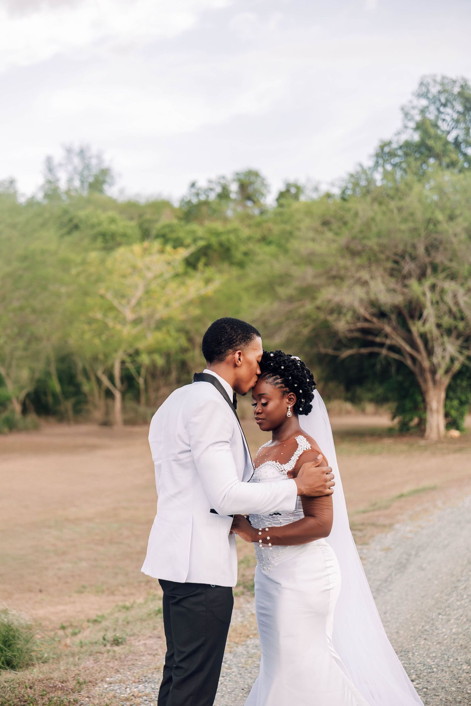 Black bride and groom sharing a tender kiss on a dirt path, bride in white mermaid gown with veil, surrounded by trees.