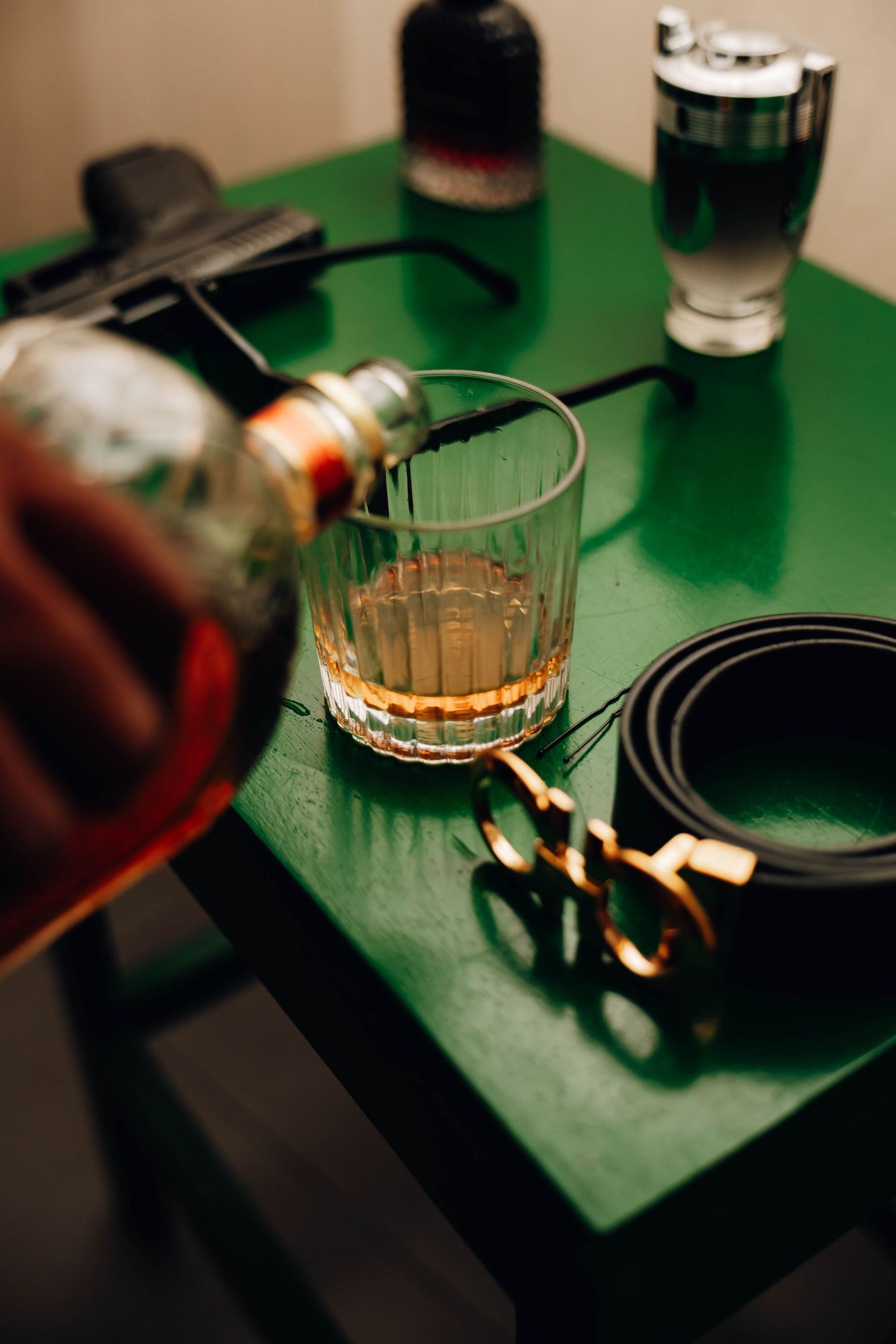 Whiskey being poured into glass beside leather belt and cologne on green table for groom prep.