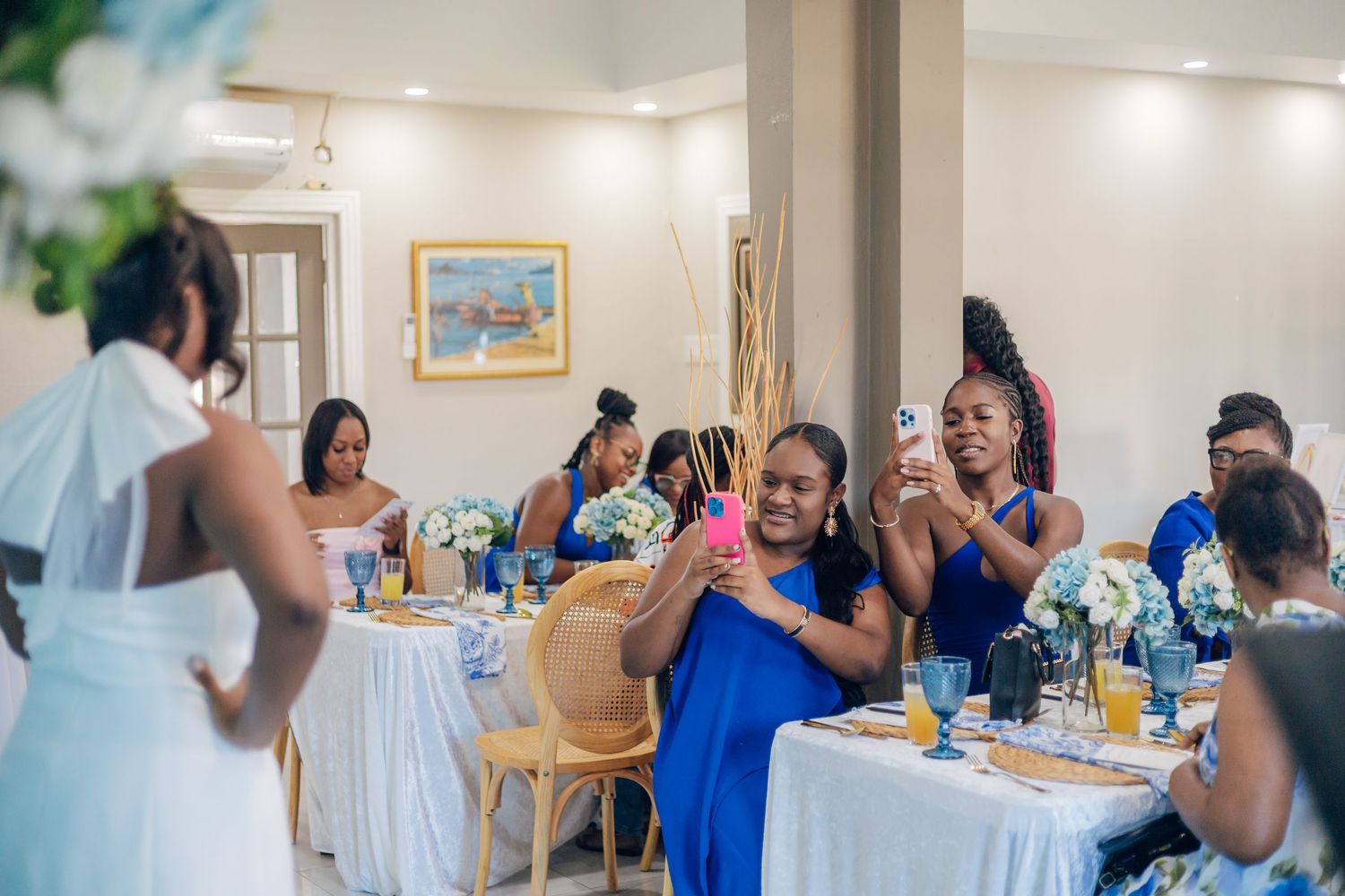 Bridesmaids in blue dresses celebrate at a wedding reception, smiling and taking photos with smartphones at decorated tables.