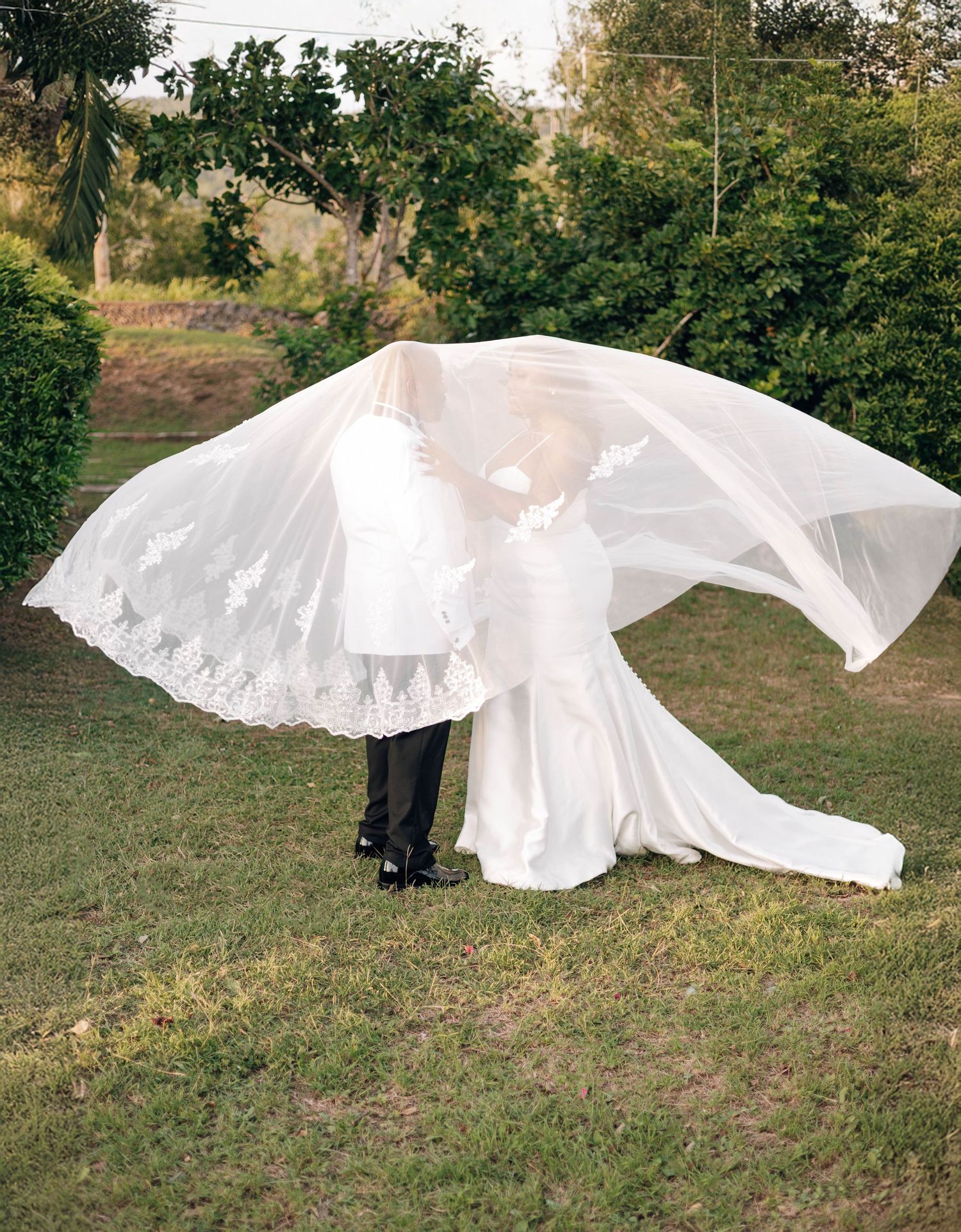Bride and groom kissing under a flowing lace-trimmed wedding veil caught by the wind in a green garden.