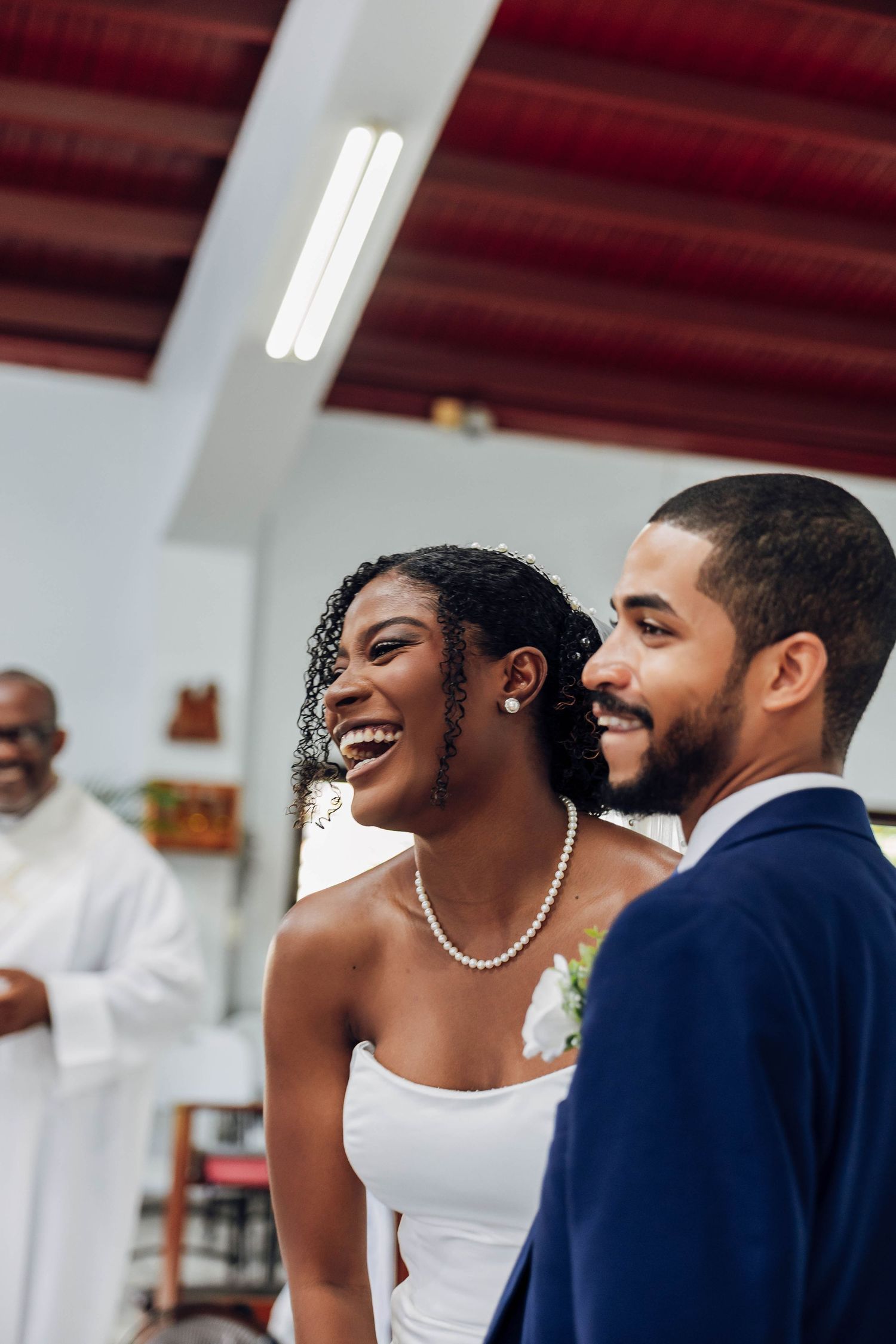 Joyful Black bride in white dress laughing beside smiling groom in blue suit during wedding ceremony inside a church.