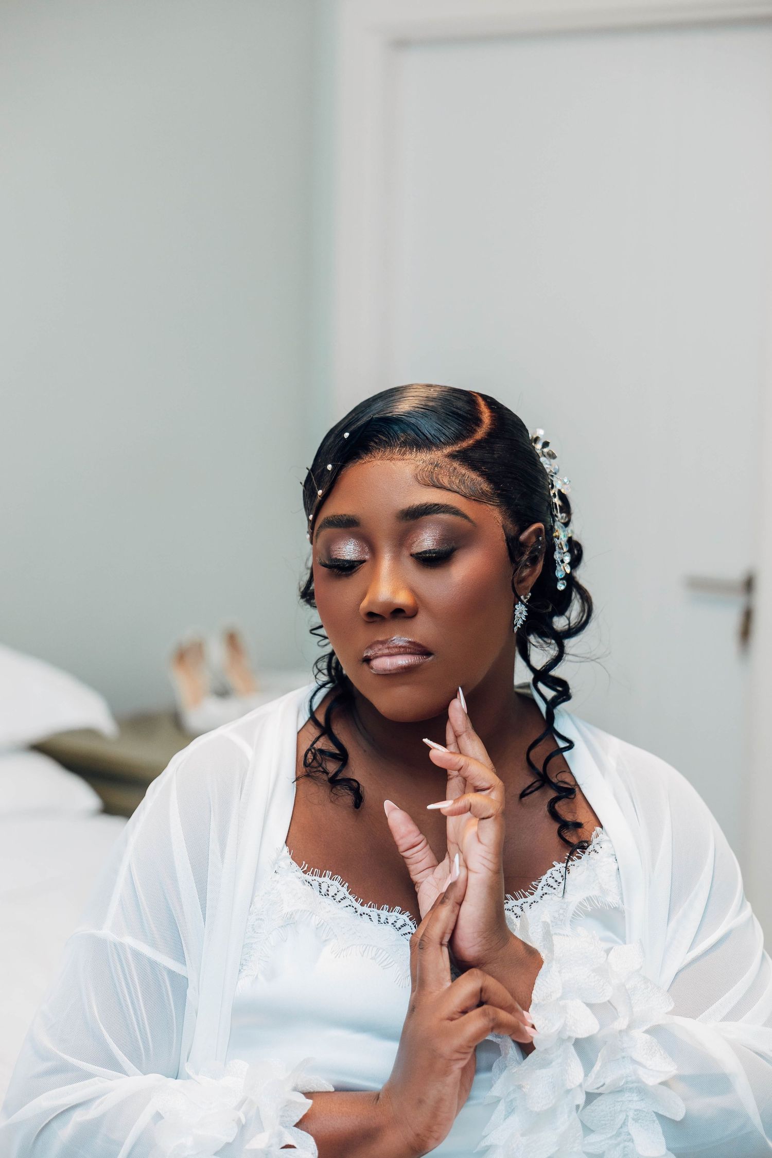 A Black bride in a white robe with curly hair adorned with crystals, eyes closed, hands clasped in a serene moment.