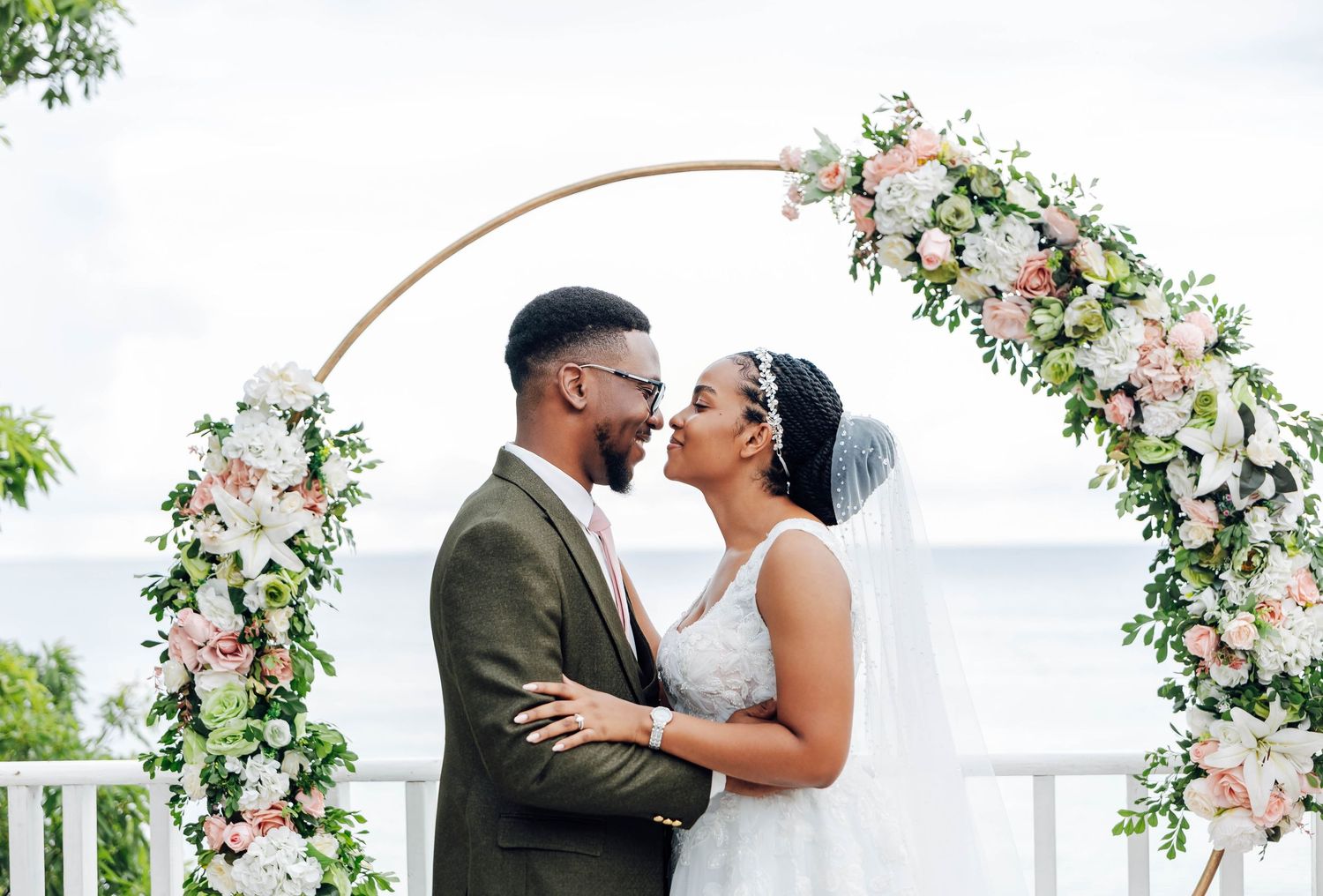 Bride and groom share a romantic moment under a floral arch with pink and white flowers at an outdoor wedding.