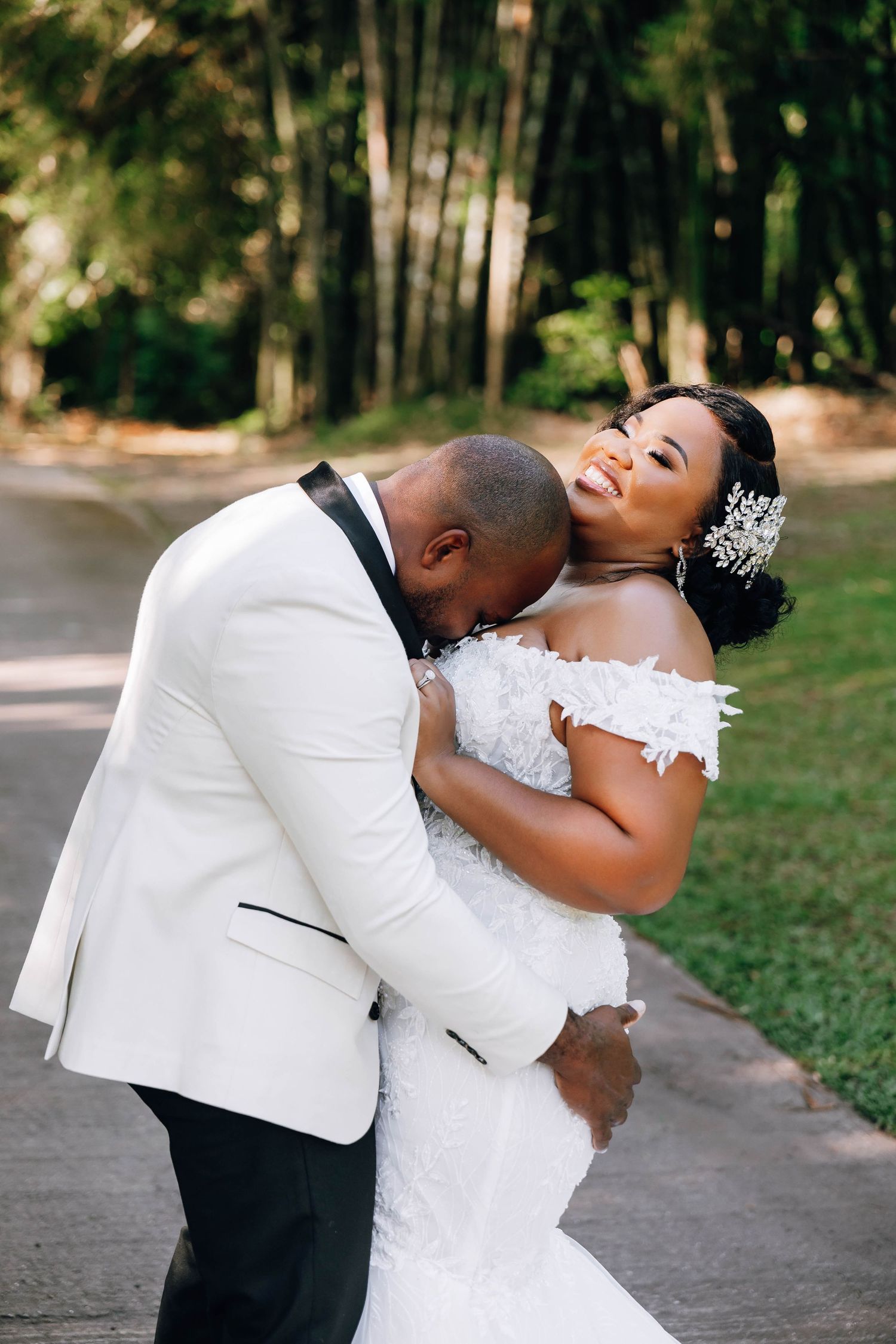 Joyful Black bride in lace gown laughing as groom in white tuxedo nuzzles her neck outdoors.