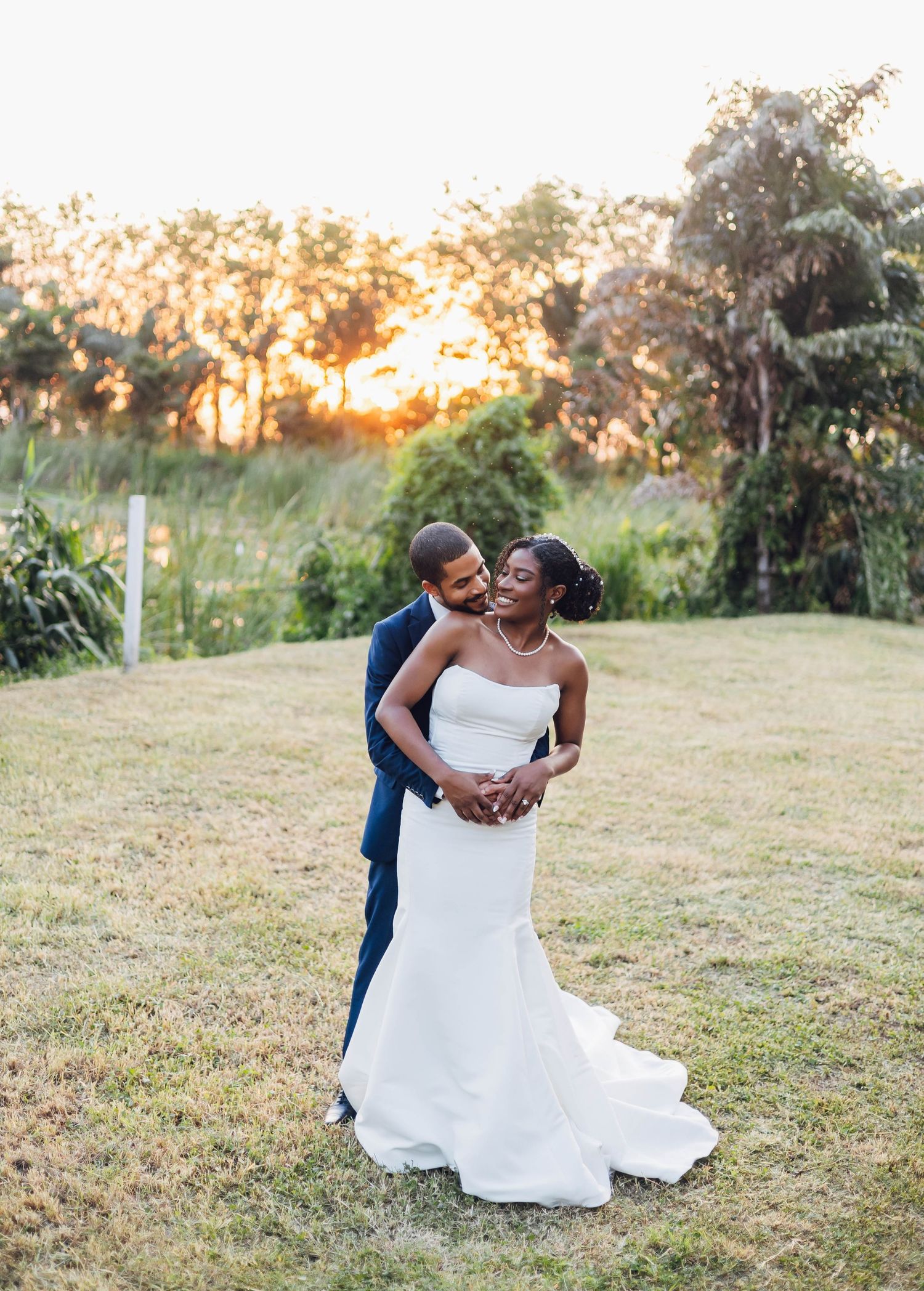 Black couple embracing at golden hour sunset in lush garden, bride in white gown and groom in navy suit.
