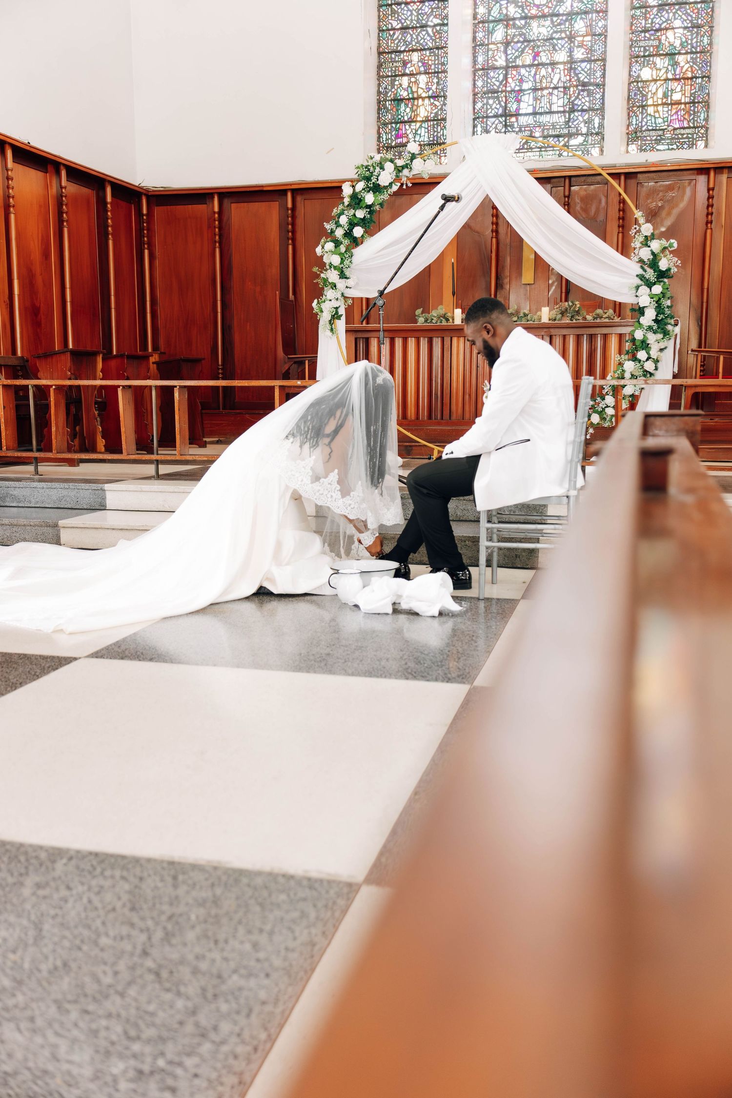 Bride and groom seated at church altar during wedding ceremony, adorned with floral arch and stained glass windows.