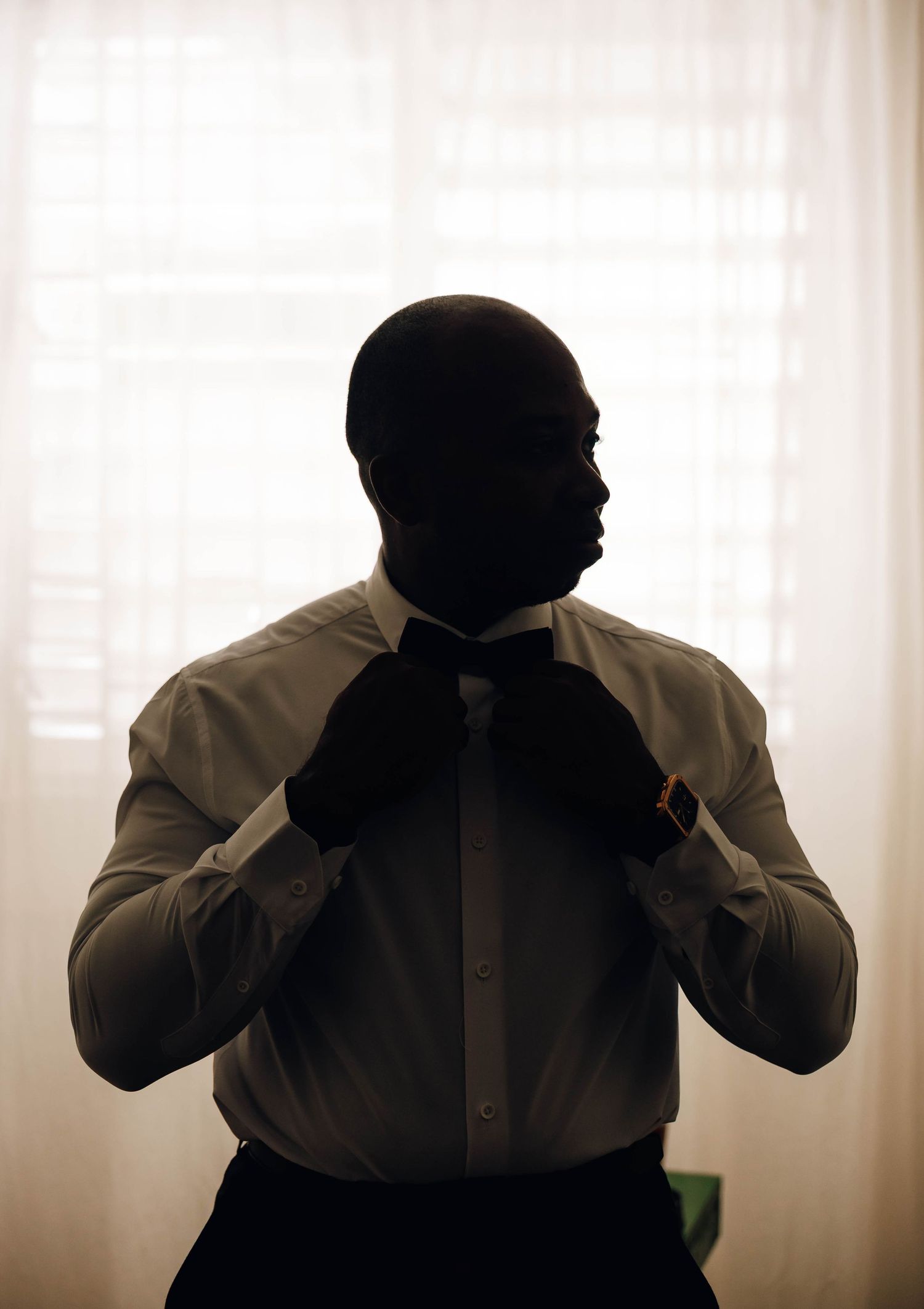 A silhouette of a groom adjusting his tie while standing in front of a bright window before his wedding.