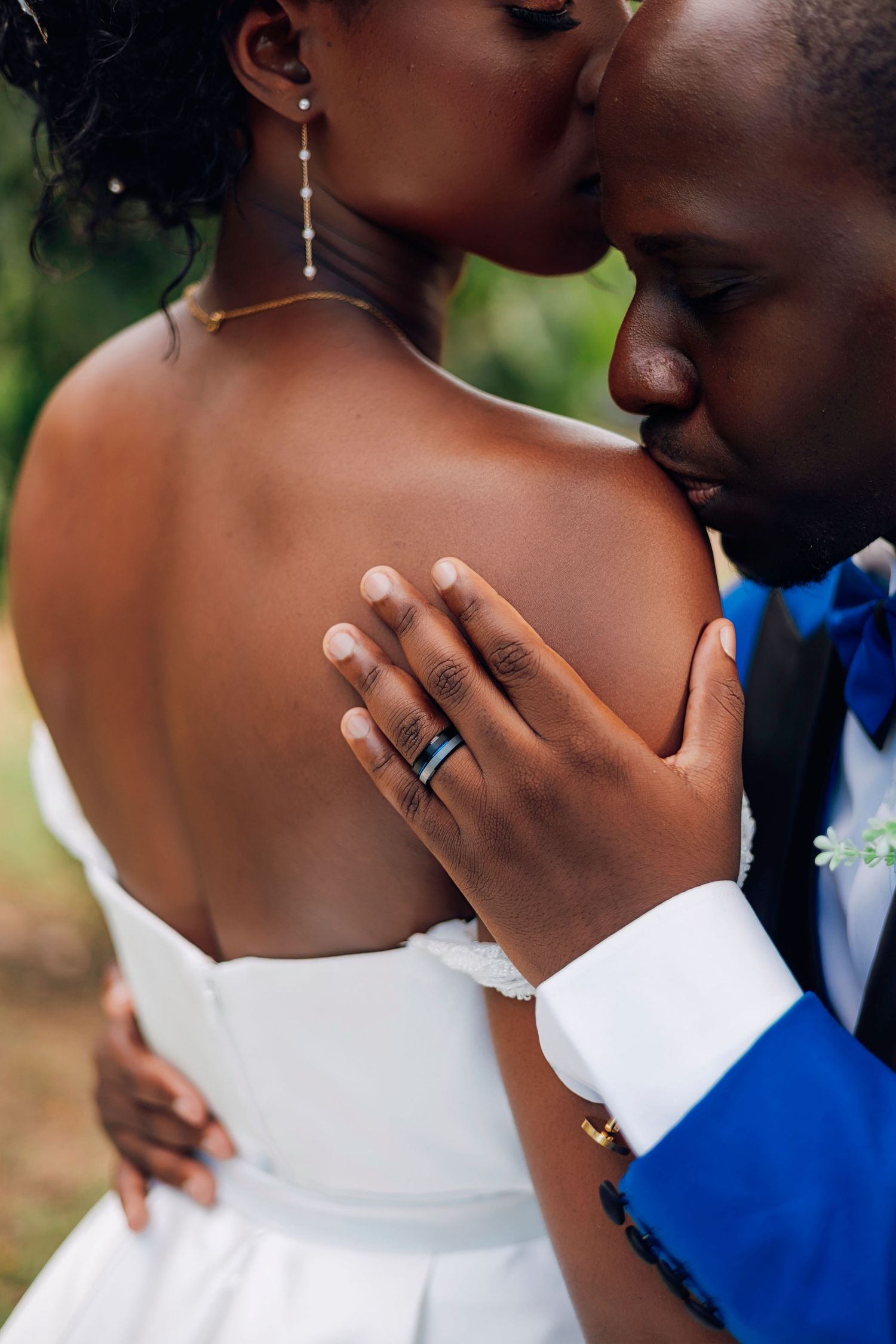 Close-up of Black groom in blue suit placing hand on bride's bare back, showcasing wedding ring, sharing an intimate moment.