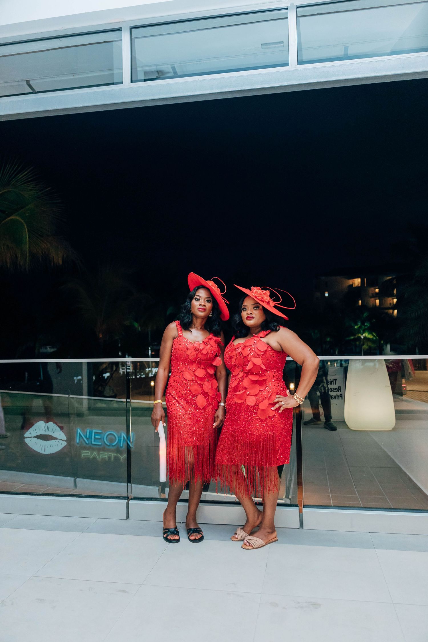 Two women in matching red lace dresses and red hats pose together on a balcony at a Neon party event.