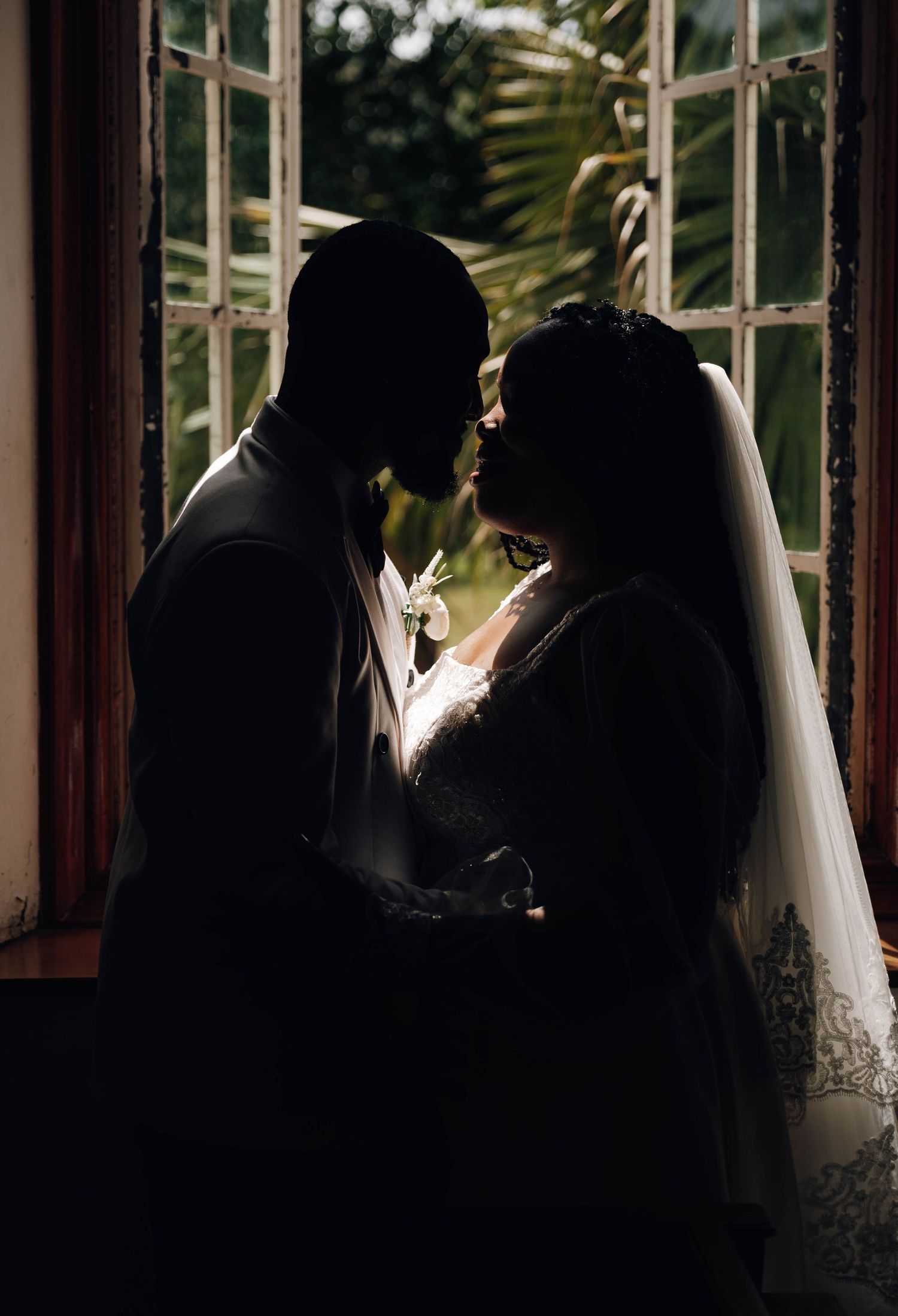 Silhouette of bride and groom sharing intimate moment by window, wedding veil and boutonniere visible in romantic backlit photo.