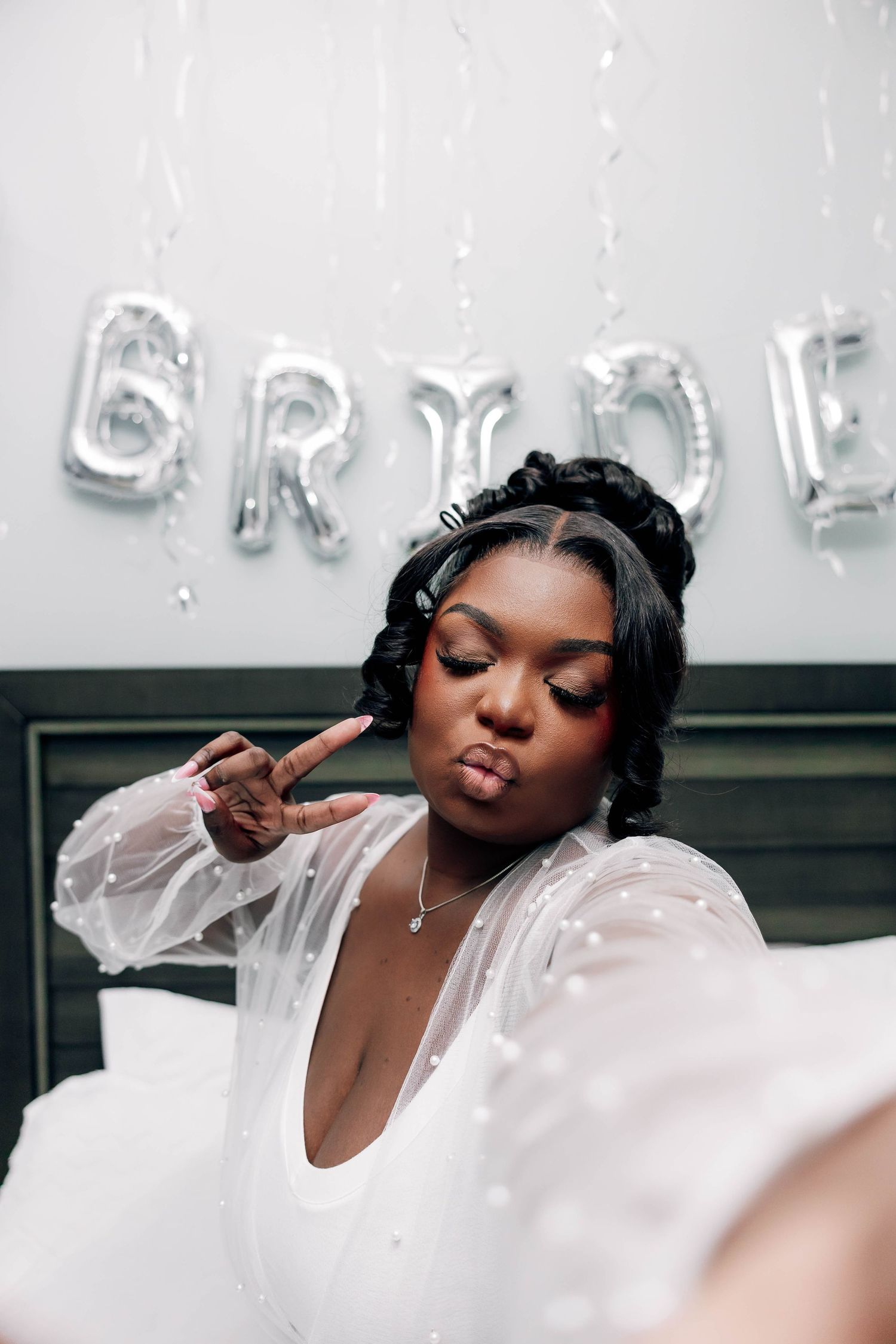 A bride in a white robe poses for a selfie in front of silver BRIDE balloon letters on her wedding day.
