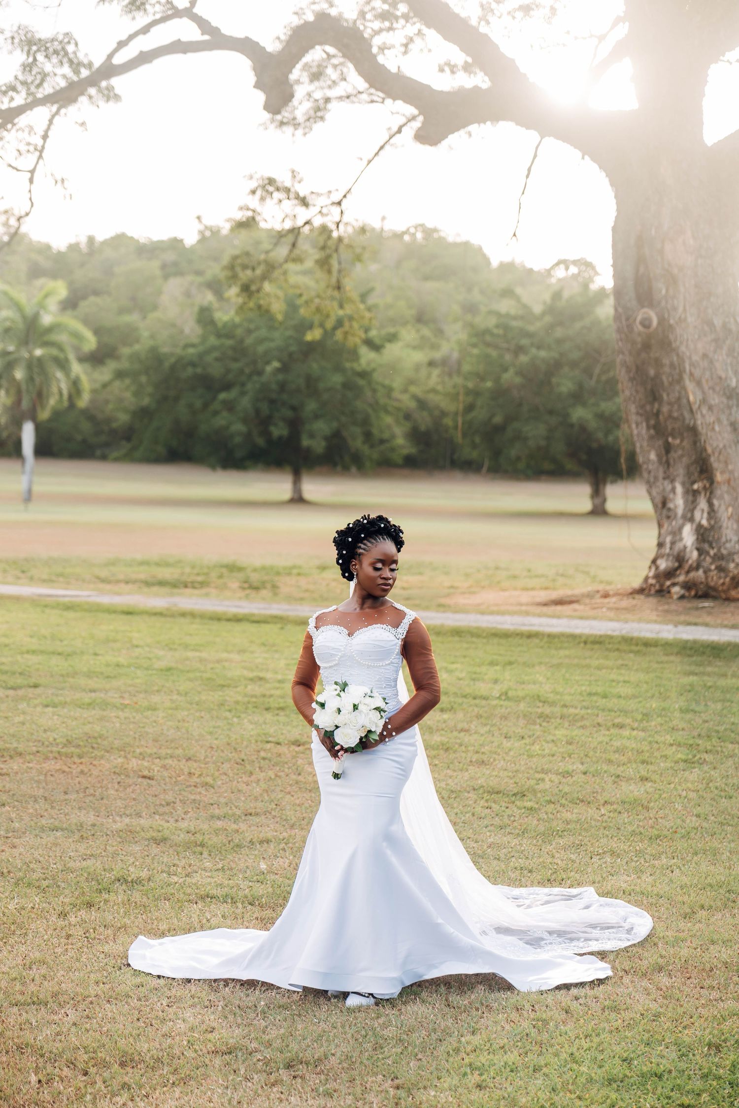 Elegant Black bride in white mermaid wedding gown with long train holding white bouquet, posing under a large tree at sunset.