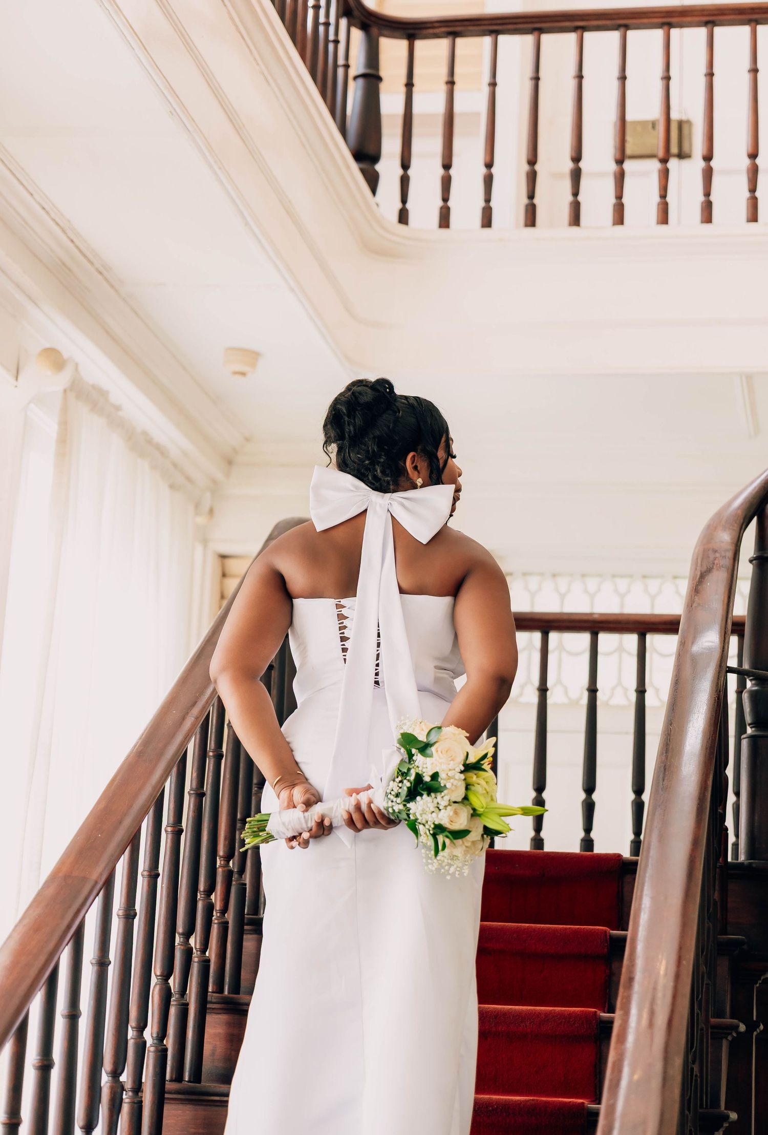 Bride in white gown with bow detail holds bouquet on red-carpeted staircase, viewed from behind.