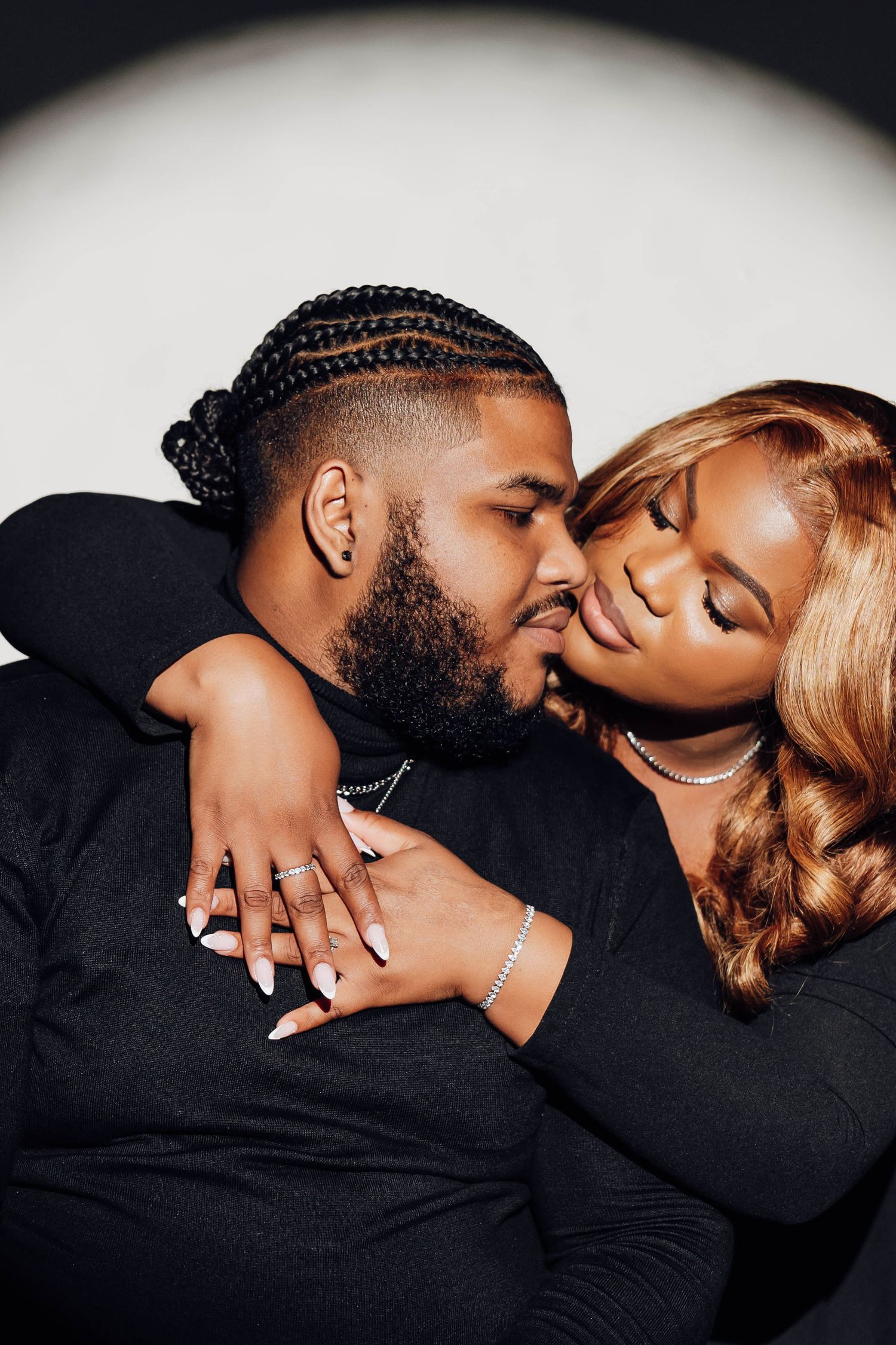 A couple in black outfits share an intimate moment, the man kissing the woman's cheek as she leans close.