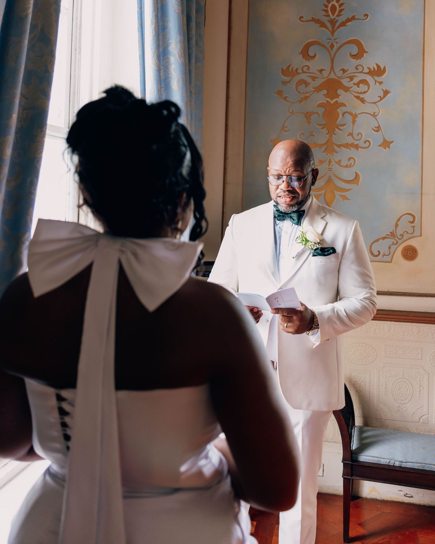 Groom in white suit reads vows to bride in backless wedding dress with bow at elegant venue.