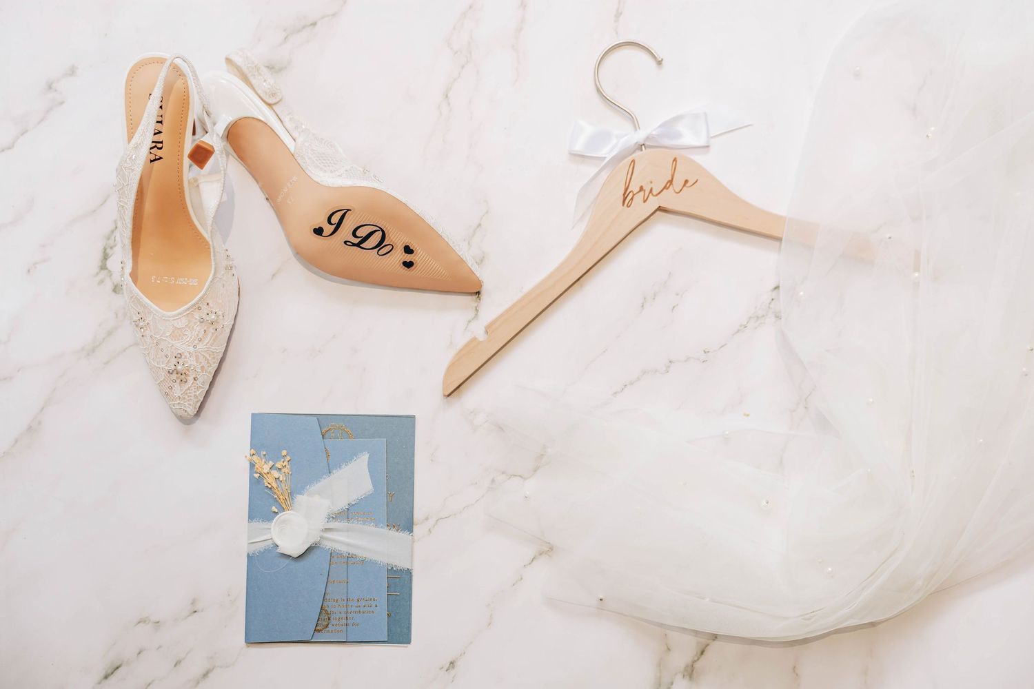 Wedding flat lay with bridal heels, wooden bride hanger, and blue invitation on marble surface.