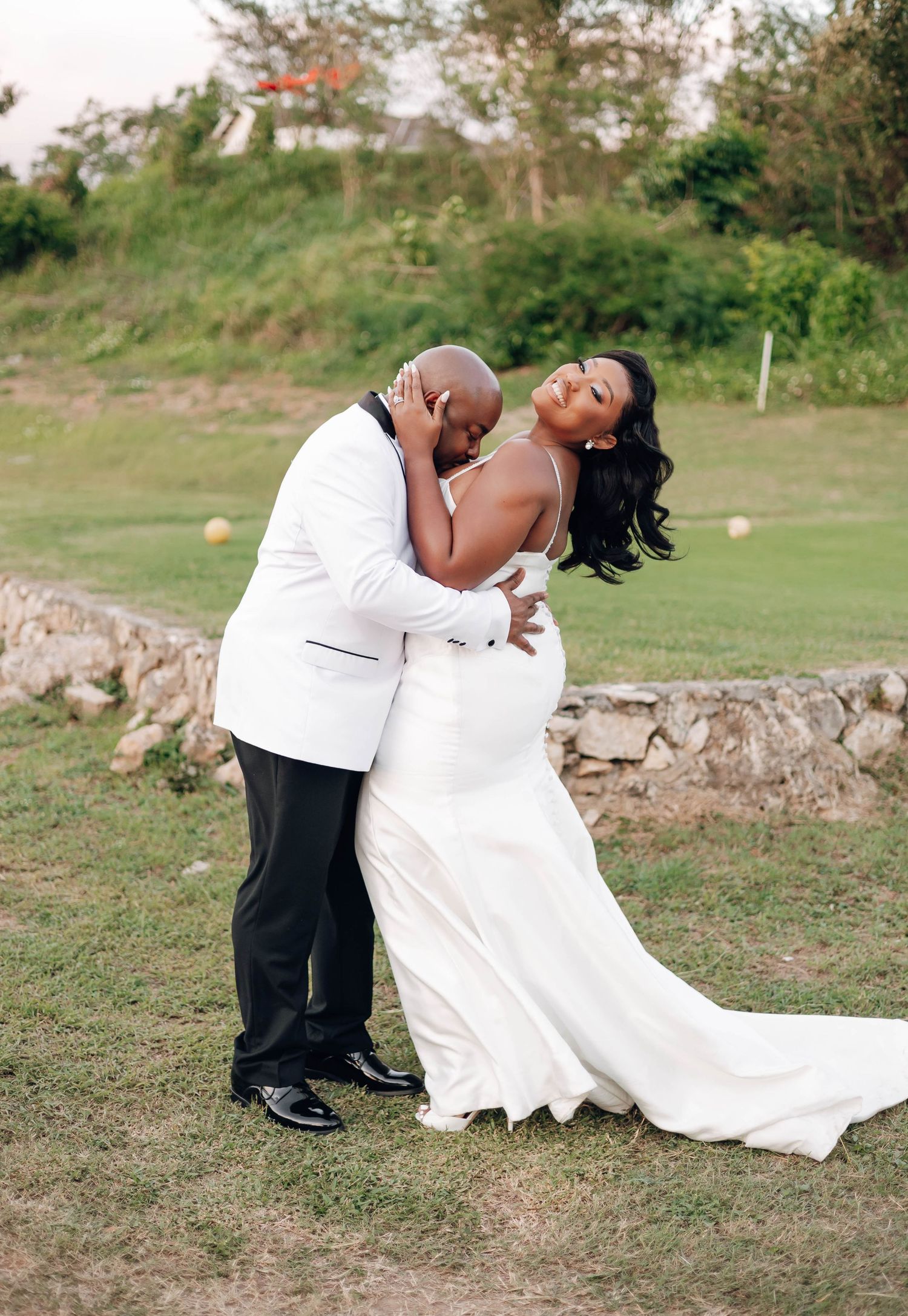 A couple shares a romantic kiss outdoors, bride in white gown and groom in white tuxedo jacket on lush green grounds.