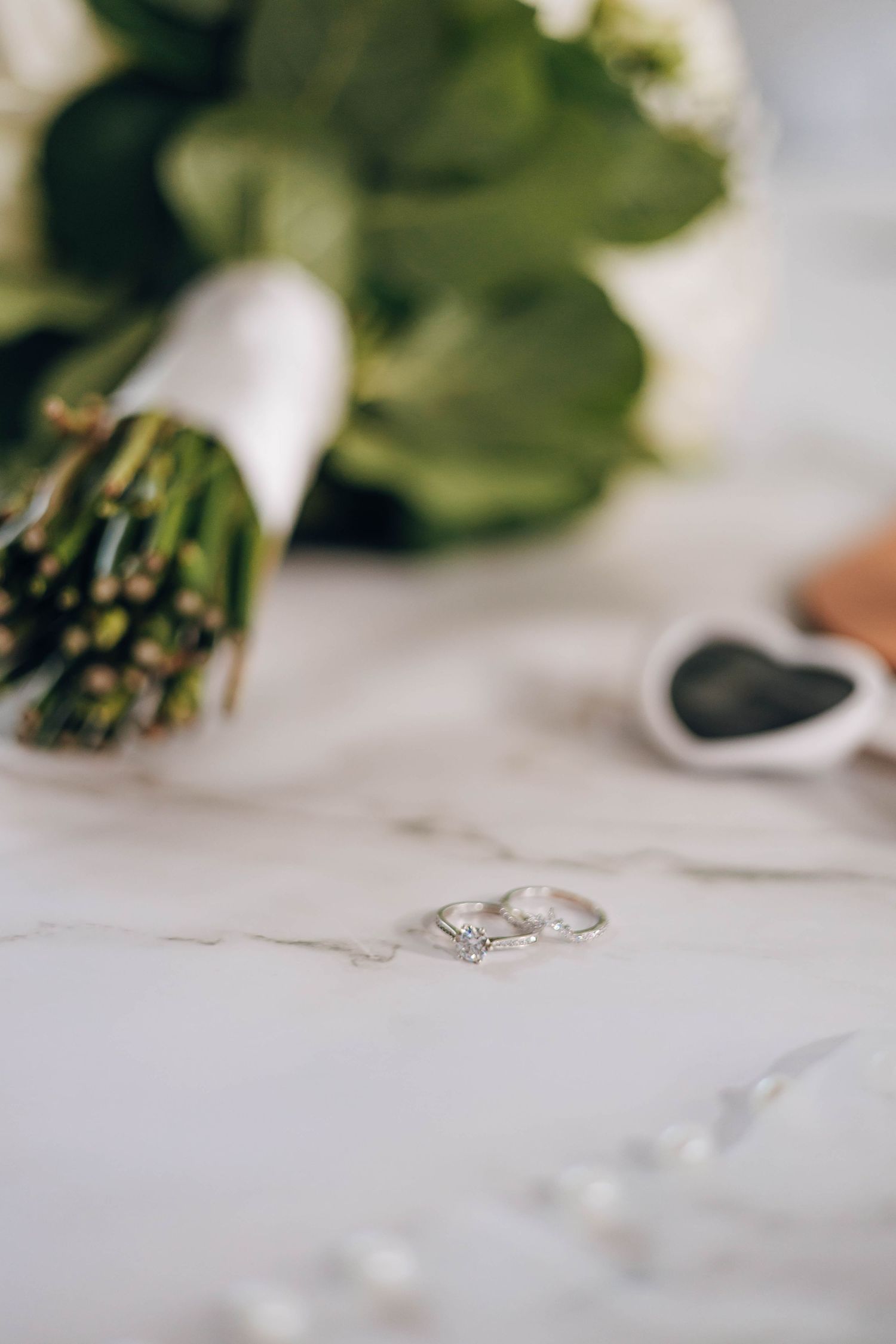 Two silver diamond wedding rings rest on a marble surface beside a white floral bouquet and heart-shaped box.