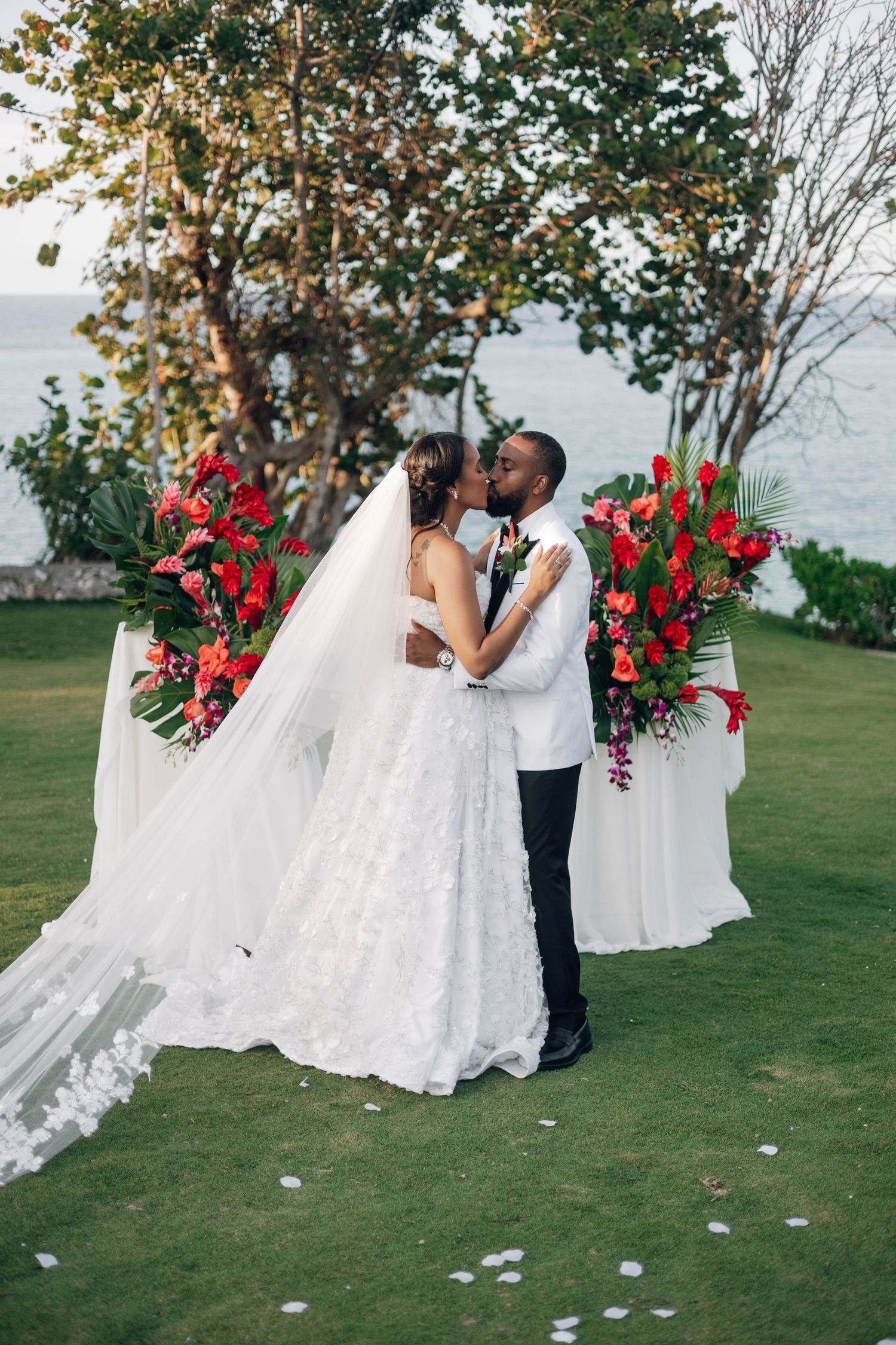 Bride and groom share first kiss at outdoor wedding altar with red tropical floral arrangements by the ocean.