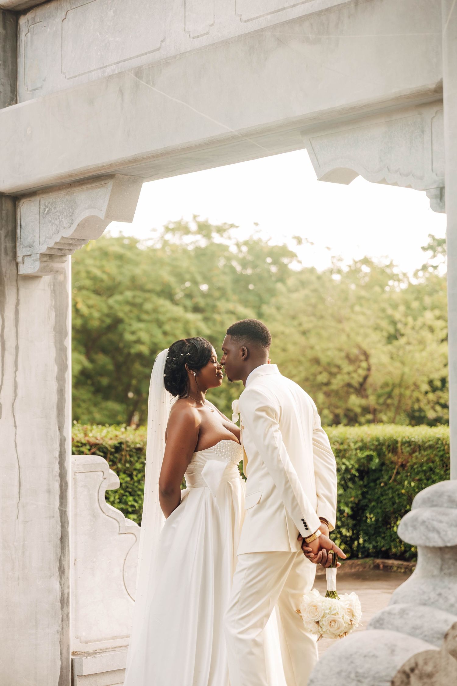 A bride and groom share a romantic moment under a stone arch surrounded by lush greenery on their wedding day.