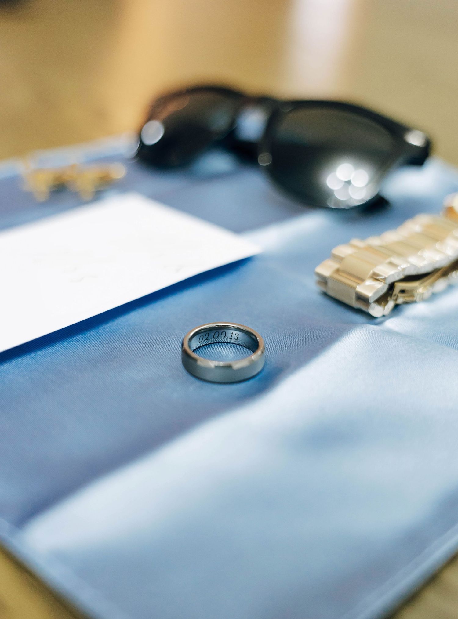 Silver wedding band ring displayed on blue fabric with sunglasses and cufflinks in background.