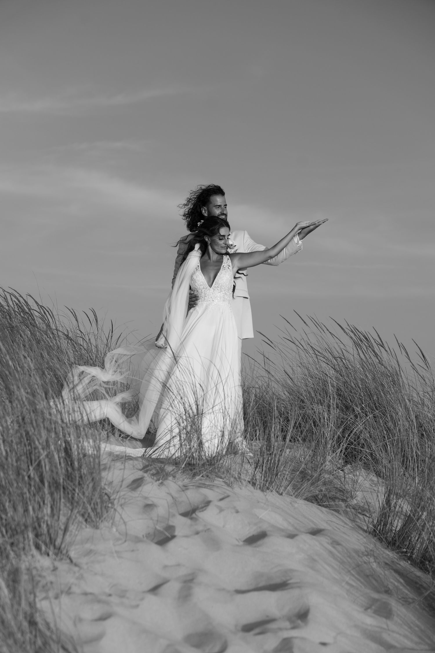 Artistic black and white portrait of a bride in flowing dress standing among beach grass.