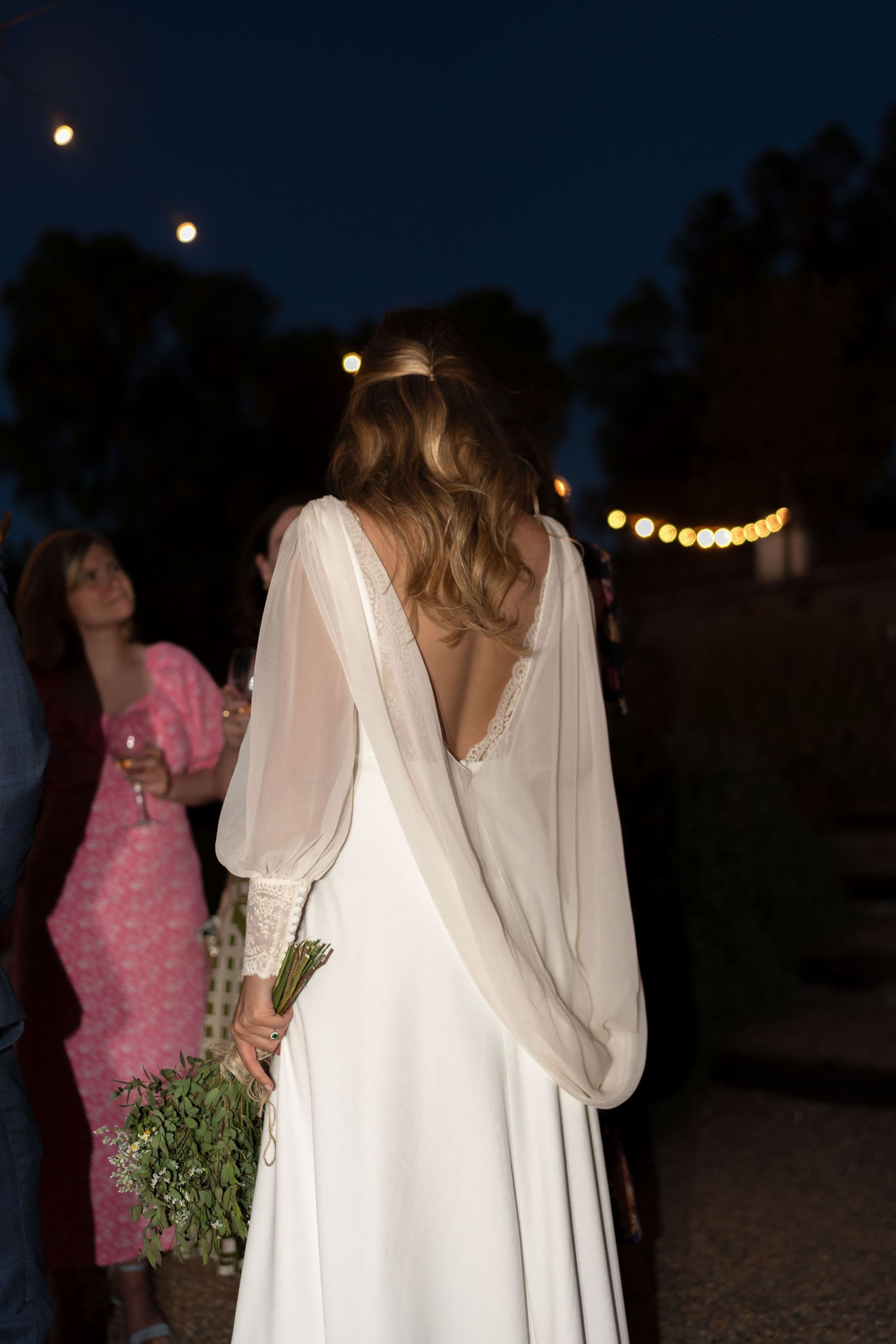 Evening wedding scene with string lights and a bride in a flowing white dress with an open back.