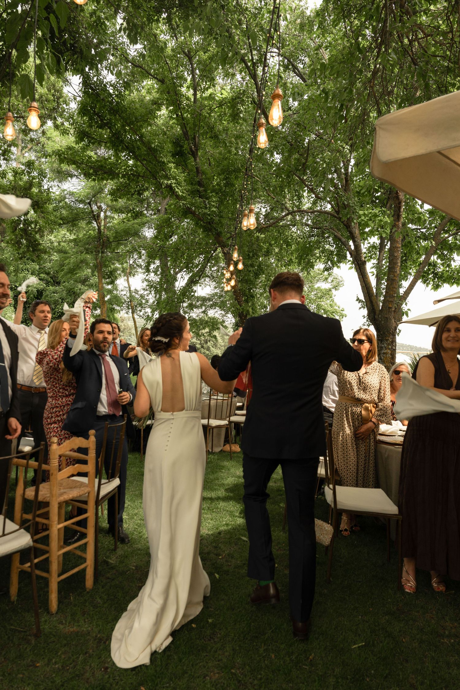 Outdoor wedding reception under trees with string lights and guests mingling.