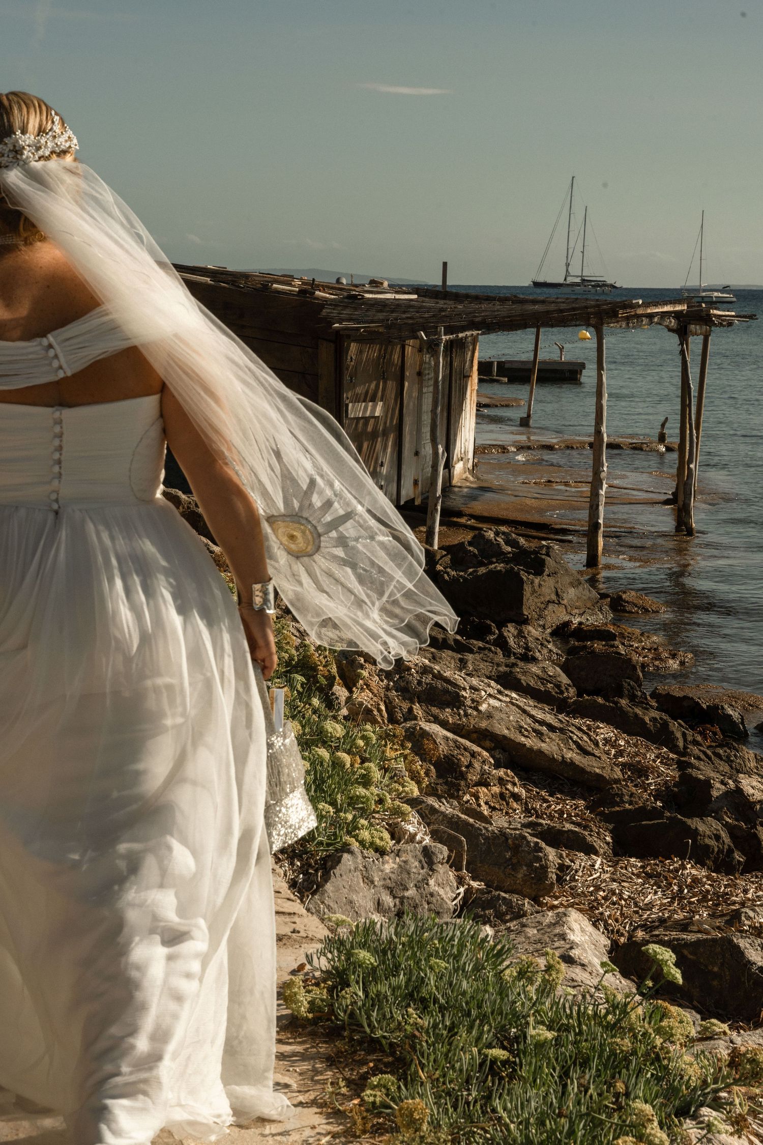 Wedding dress flowing in breeze near coastal dock and rocks.