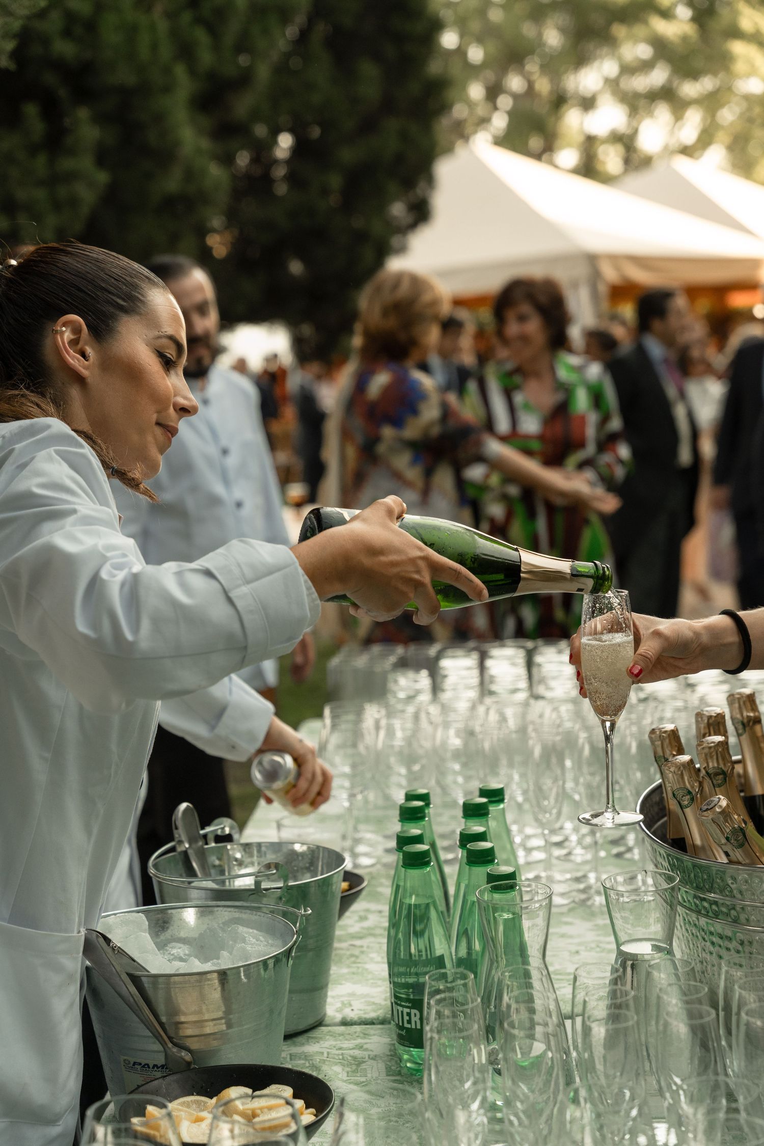 Bartender in white uniform pouring drinks at an outdoor event with rows of glasses on table.