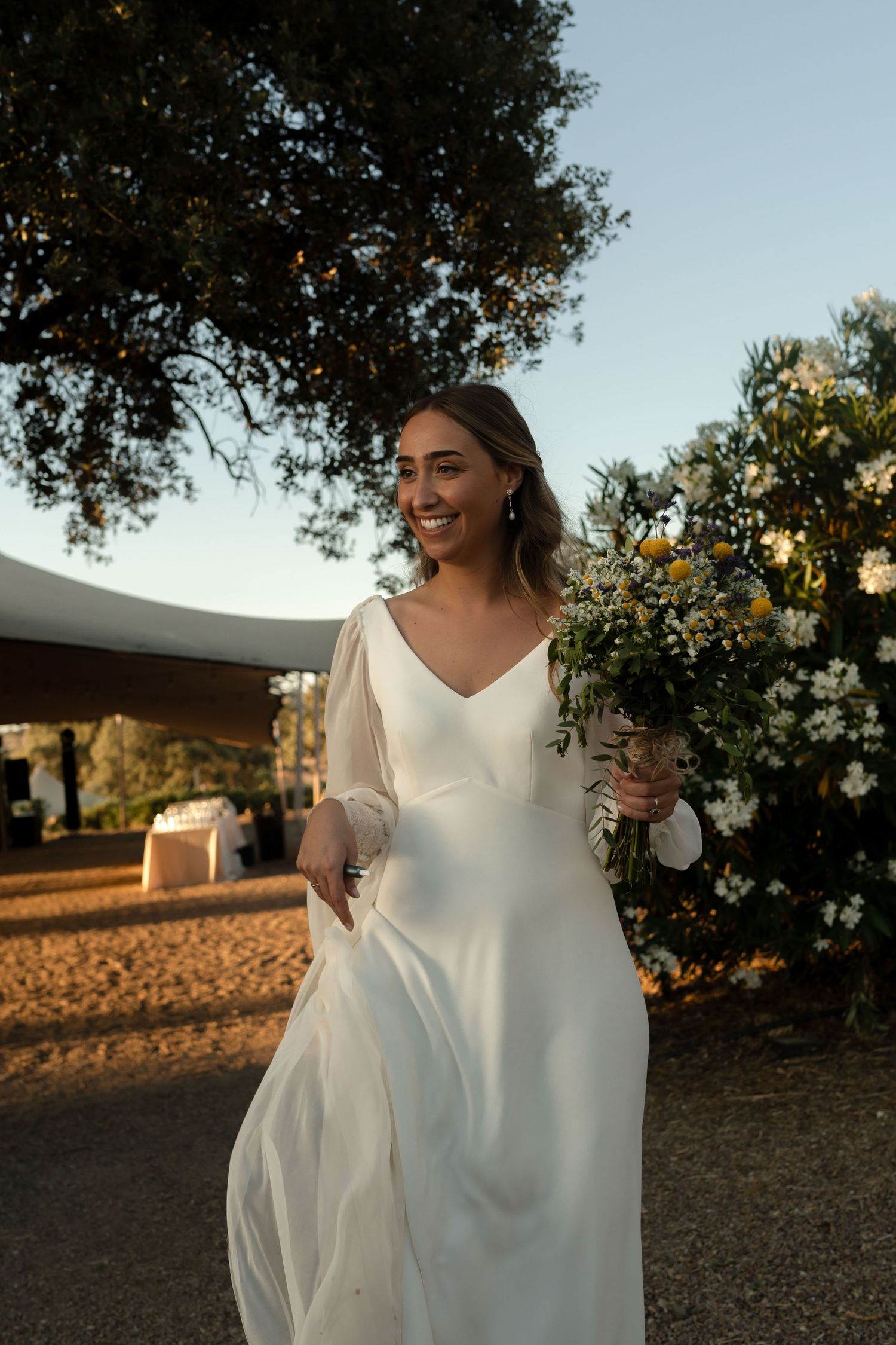 A bride holds her dress and bouquet while walking through an outdoor venue at sunset.