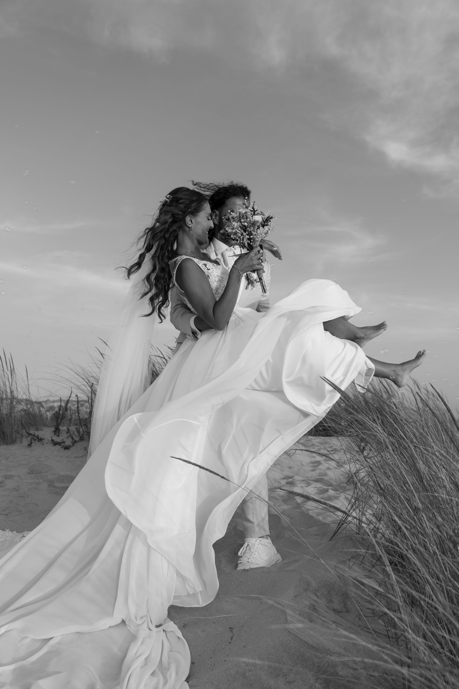 Dramatic black and white photo of a bride with flowing dress in coastal dunes.