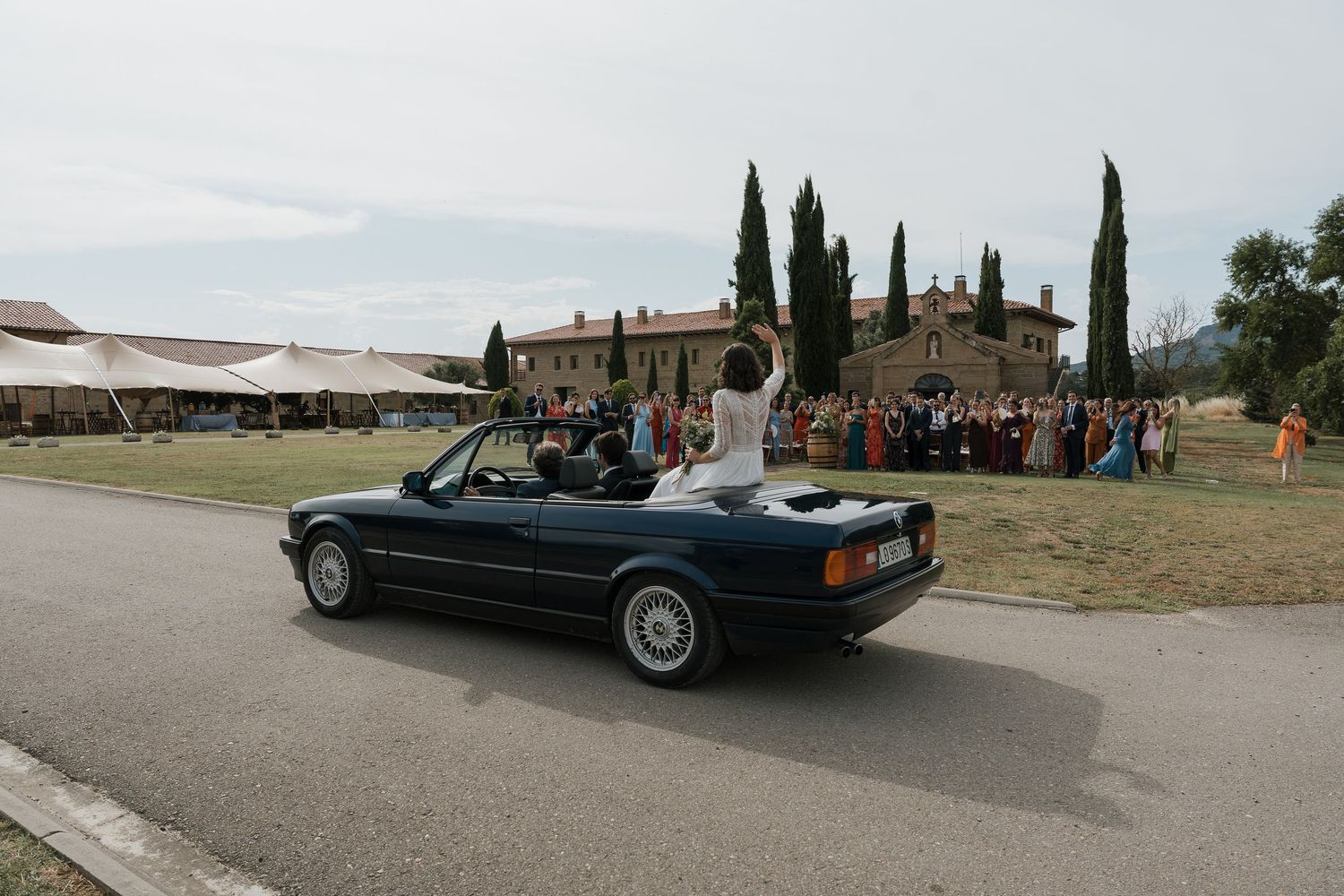 Vintage BMW convertible carrying newlyweds past Tuscan villa with celebrating guests.