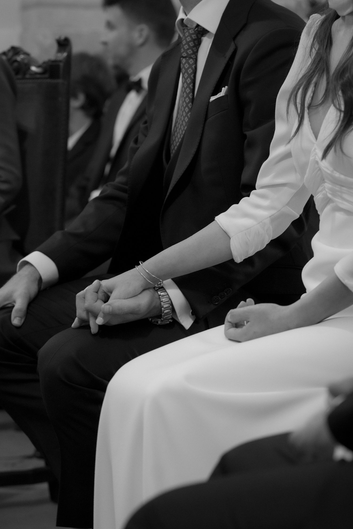 Wedding guests seated in formal attire on white chairs during an indoor ceremony.